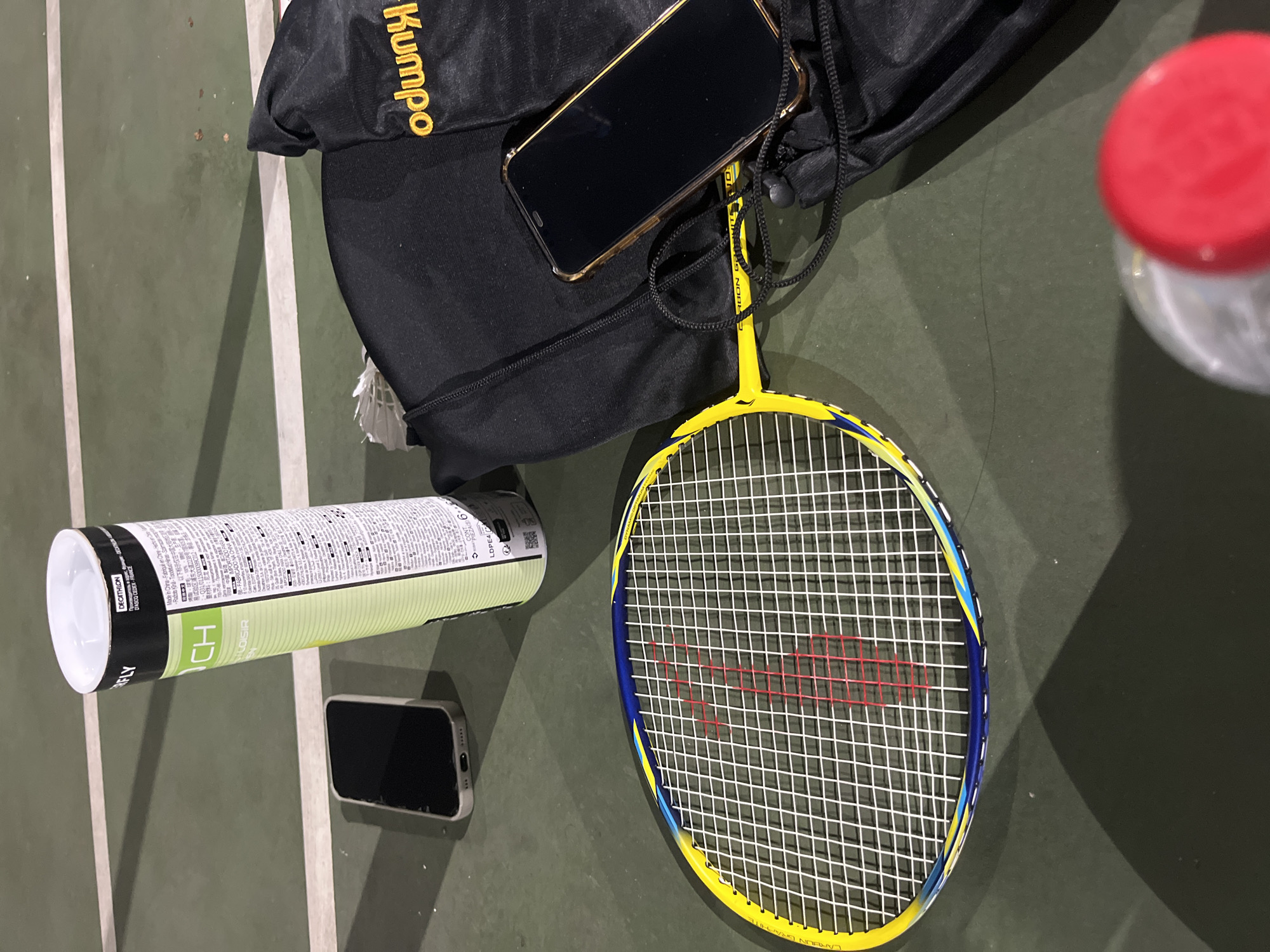 A badminton racket, a can of shuttlecocks, two smartphones, and a black bag are laid out on a green court, possibly in Yuzhong District, China. The badminton racket is yellow with blue and yellow accents on the frame. The can of shuttlecocks is tall and cylindrical, with text and branding visible. One smartphone is placed to the left of the can, and another, larger smartphone with a golden frame, rests near the black bag. The black bag has yellow lettering that reads "odumpy." A partially visible red bottle cap is in the foreground, suggesting a drink is present. The court appears to be outdoors, with faint white lines marking boundaries.