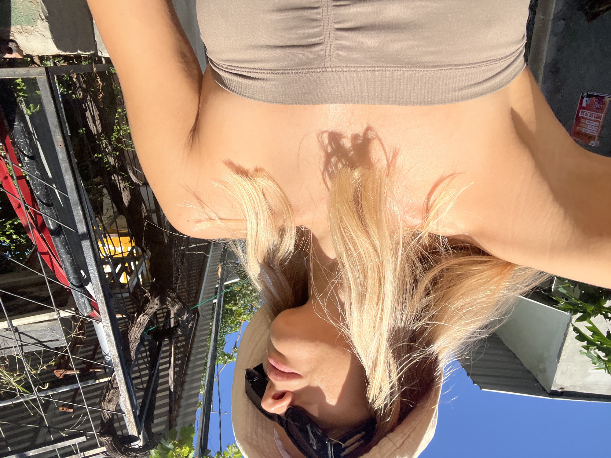 The image shows a close-up, low-angle view of a blonde woman outdoors, wearing a tan bucket hat and dark sunglasses. She is looking upwards and to the right, with her left arm raised and partially visible. The woman is wearing a ruched, strapless top in a taupe color. Sunlight highlights her skin, casting shadows on her chest.
In the background, to the left, is a portion of a grey building with a corrugated metal roof. To the right, a black metal fence encloses an area with plants and foliage, including vines that appear to be growing on the fence structure. A bright yellow object is visible through the fence. A red object is also partially visible. A sign with Vietnamese text and imagery is affixed to the grey building on the left. The sky is a clear, bright blue. The scene appears to be in an outdoor urban or semi-urban environment, possibly a residential area or a less developed part of the city. The presence of the sign with Vietnamese text and the style of the buildings suggest the location is in Vietnam. Given the context of Nha Trang Ward, Vietnam, this could be a street view or a yard area.