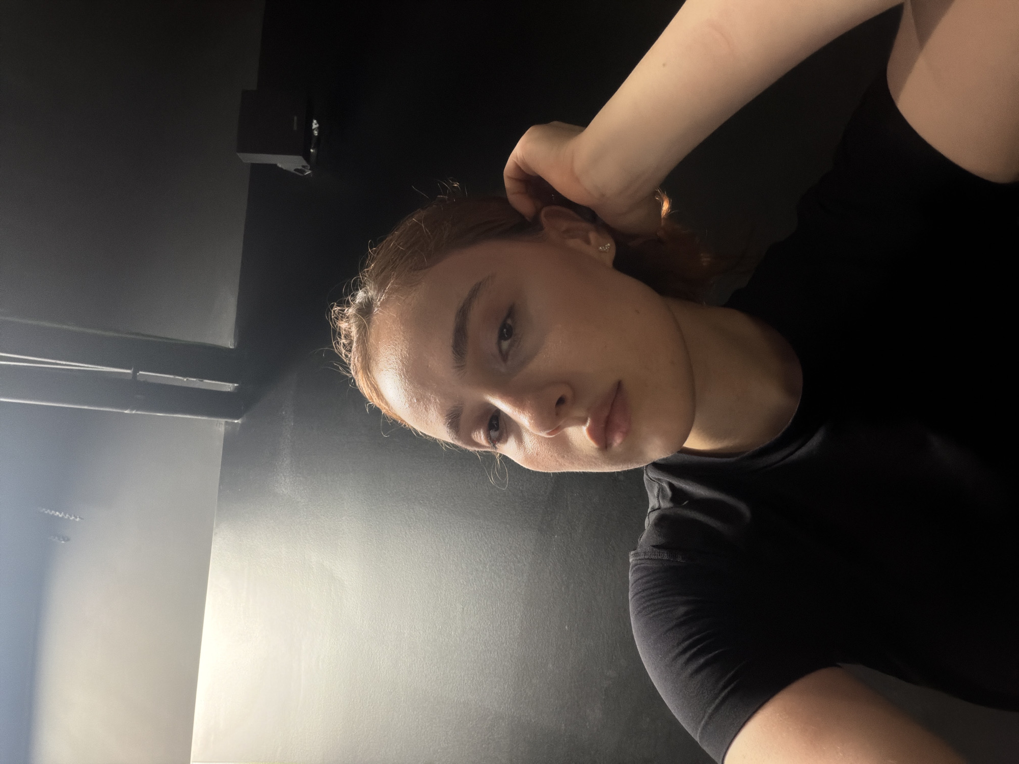 The media file shows a young woman with auburn hair, wearing a black t-shirt. She is in a dimly lit indoor setting, possibly a room or studio, with dark walls and ceiling. One part of the wall is illuminated by a bright light source, creating a contrast. The woman is looking towards the camera, with her right hand touching her hair. Her expression is neutral. The image is oriented sideways. No text is visible. The setting appears to be modern and minimalist. There are no obvious clues about the specific time of day or weather conditions.