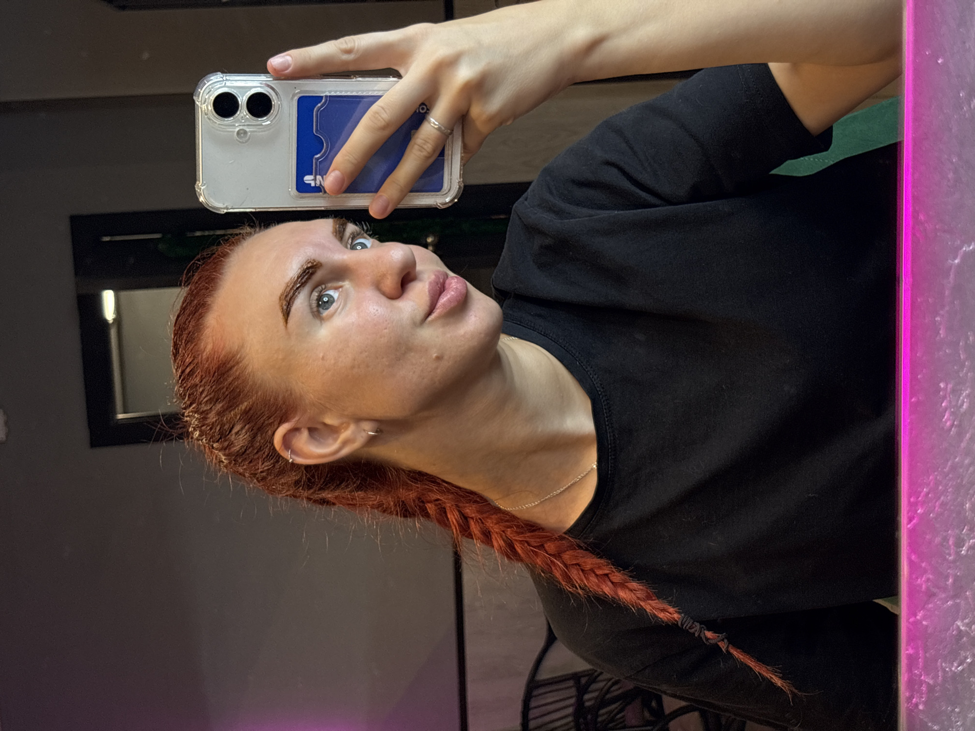 A woman with red hair styled in a braid takes a mirror selfie. She is wearing a black t-shirt. Her phone has a clear case with a blue cardholder. The background appears to be an indoor setting with dark walls and a hint of purple lighting. The image does not provide any specific details to determine its location within Rostov-on-Don, Russia.