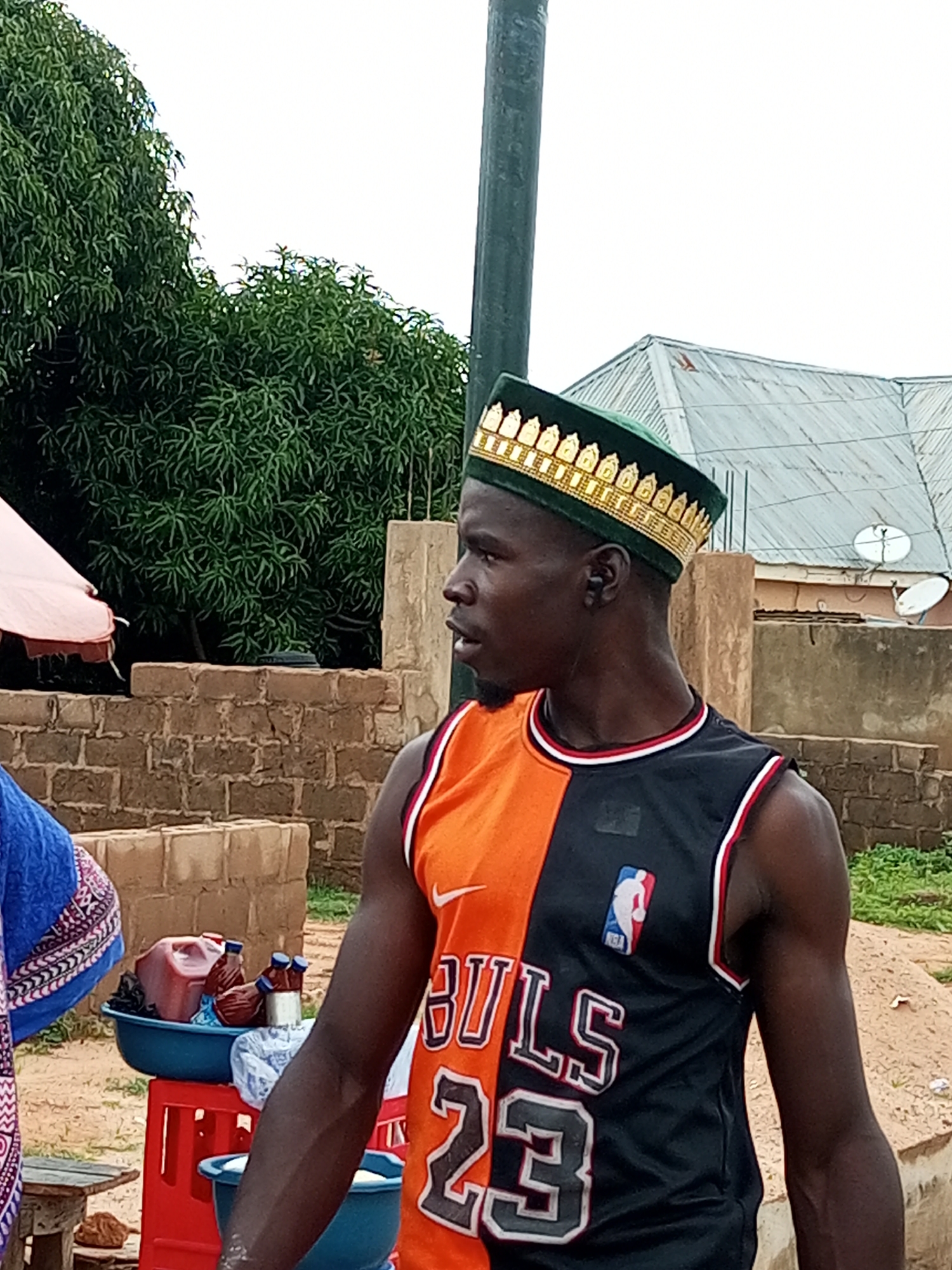 In Tempure, Nigeria, a young man stands in profile to the right. He wears a green cap with gold embellishments and a black and orange sleeveless jersey that reads "BULLS 23" in white lettering. The jersey has a Nike logo and an NBA logo. He has dark skin and short, dark hair. The man appears to be looking at something out of frame to the right.
Behind him, a brick wall forms the backdrop. To the left of the man, a large tree with lush green leaves is visible. Further in the background, a building with a corrugated metal roof and a satellite dish can be seen. In the foreground, to the left of the man, is a portion of another person wearing a blue garment with a patterned trim. There are also two blue plastic basins on a red plastic crate, one containing a large red jug and several brown bottles, and the other appearing to be empty.