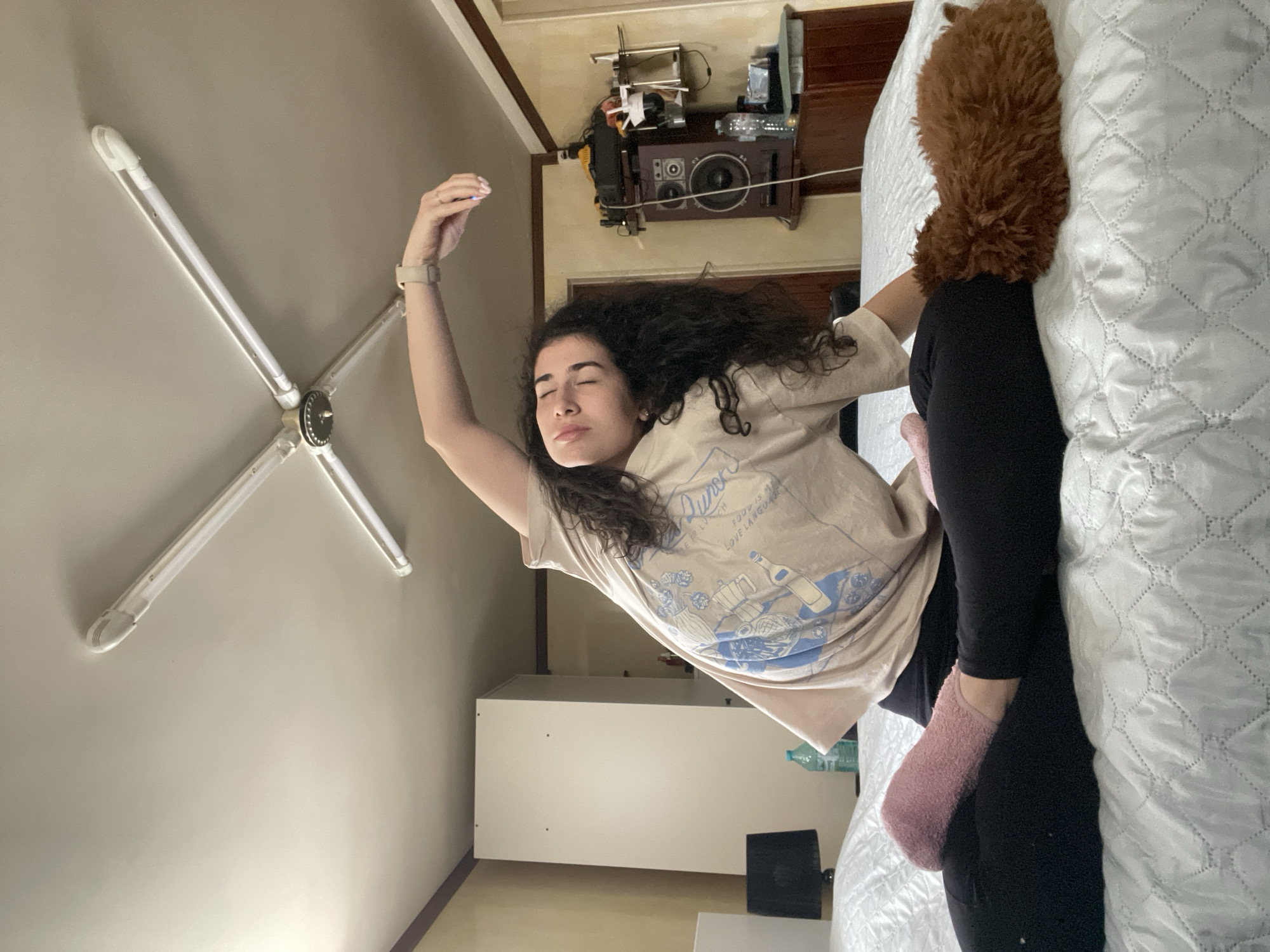 A woman with long, curly dark hair lies on a bed with a white quilted comforter. She wears a light beige t-shirt, black leggings, and pink fuzzy socks. Her left arm is raised above her head, adorned with a watch. A brown fuzzy item rests on the bed next to her. The room appears to be inside, lit by an overhead light fixture with four fluorescent bulbs. The walls are light colored. A white cabinet is visible in the background, along with a stereo system and other household items. Based on the furniture and general style of the room, it is difficult to ascertain a specific location within Sofia, Bulgaria. The scene appears to be a personal space, possibly a bedroom.
