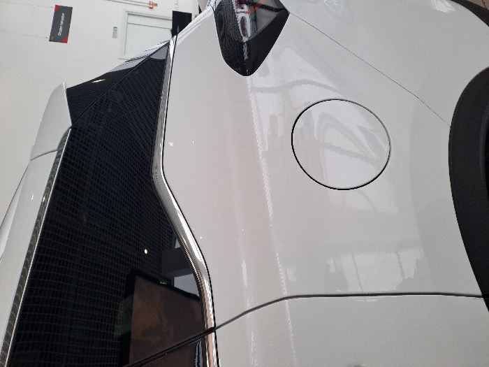 A close-up shot reveals the rear quarter panel of a white vehicle. The panel features a round fuel filler door with a defined black outline. A black tire is partially visible on the right. To the left, a dark, textured panel with a silver trim runs vertically. The scene is well-lit, highlighting the vehicle's glossy finish and design elements. A reflection on the textured panel suggests an interior or background scene.