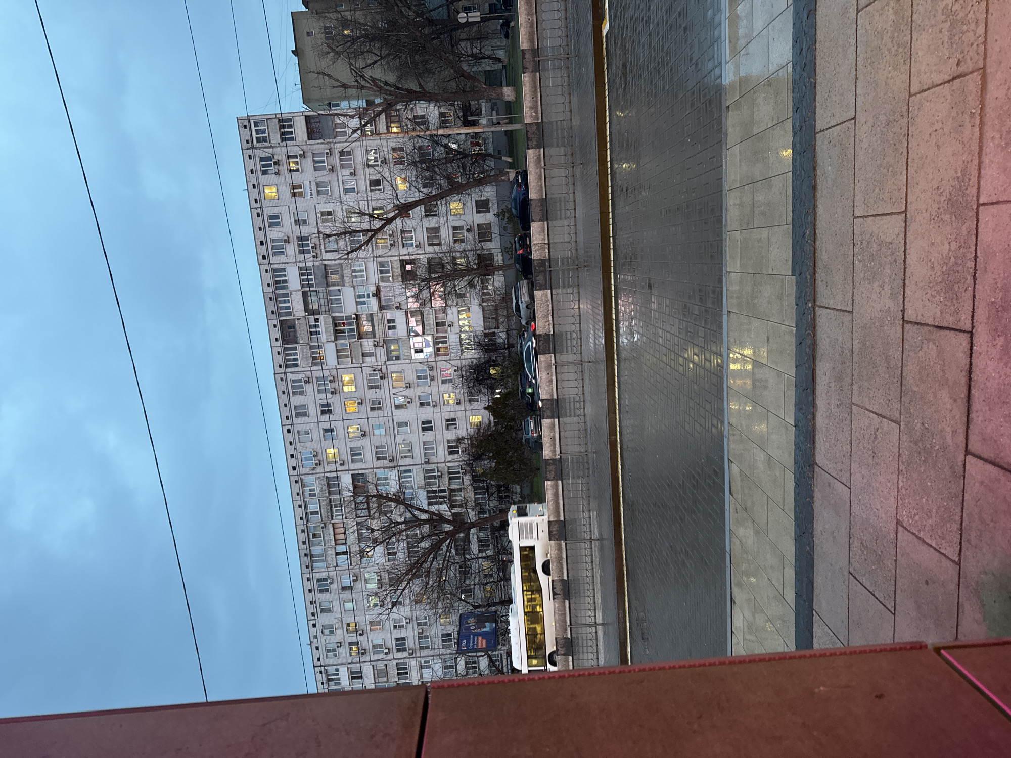 The image shows a street scene in Ol'ginskaya, Russia. A large, Soviet-era apartment building dominates the left side of the frame, with numerous lit windows suggesting it is evening. Bare trees stand between the building and the street. Cars are parked along the curb, and a white bus is visible. The street is paved with grey tiles, wet in places, reflecting light. A pedestrian walkway, composed of different colored paving stones, runs along the right side of the frame. Overhead power lines are visible in the sky. The view is from an elevated position, looking down at the street. The lower portion of the image is obscured by an out-of-focus reddish-brown structure.