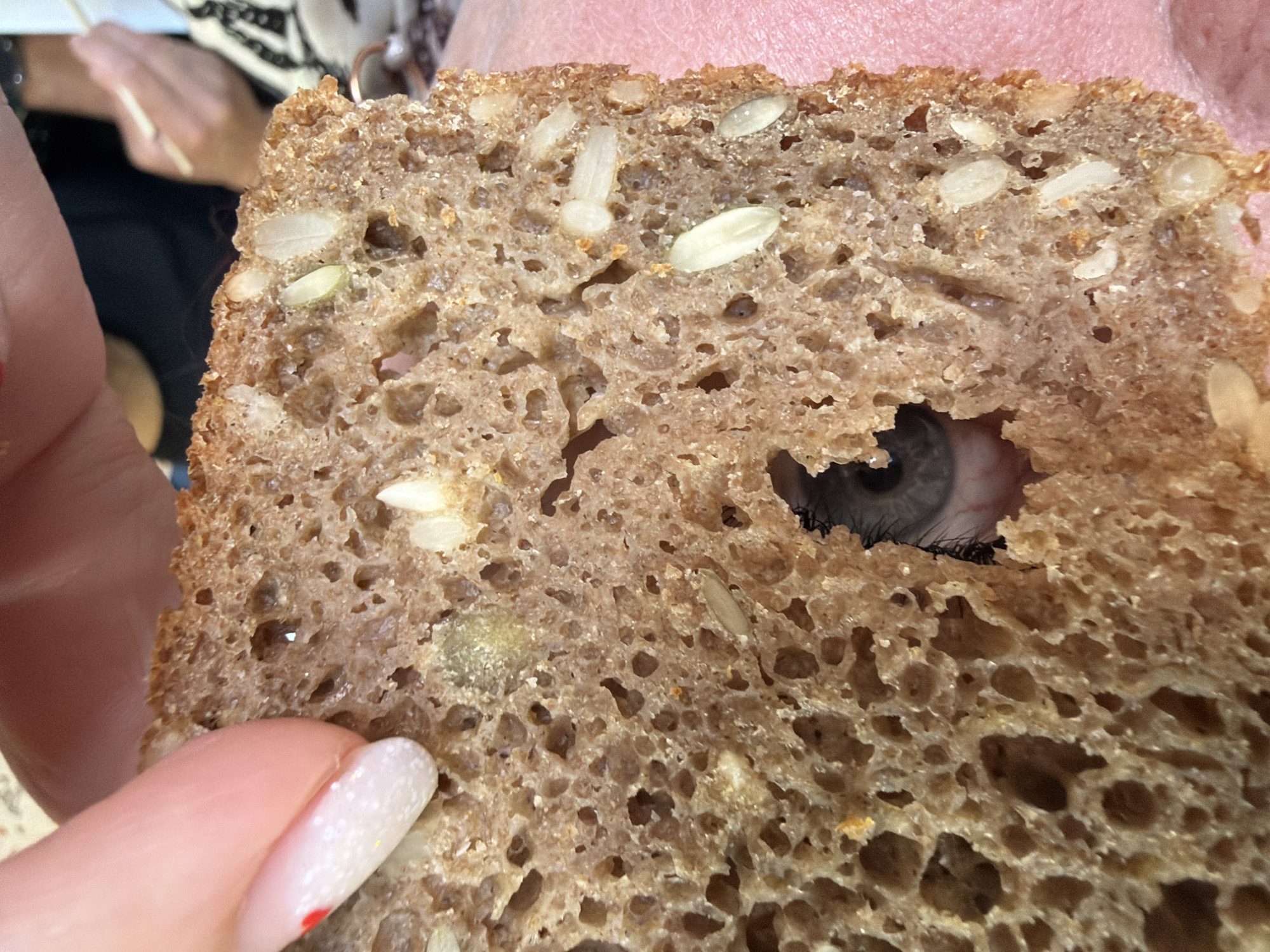 A slice of brown, seeded bread is being held up with a hole through which an eye is visible. The person holding the bread is also visible on the sides of the image. The location is not identifiable as Chaikovsky, Russia.