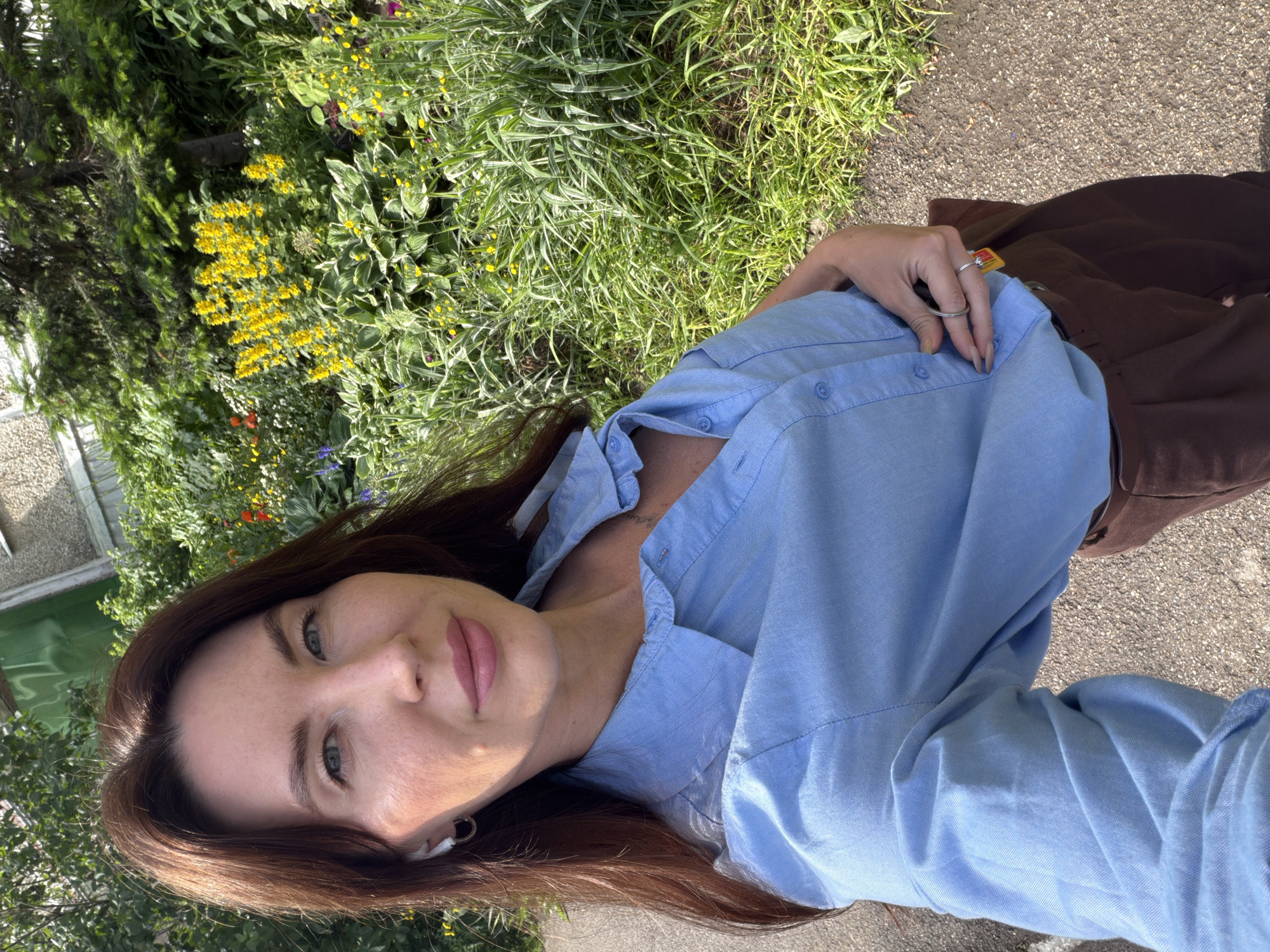 A woman takes a selfie outdoors in a city park in Perm, Russia. She has long, dark brown hair and is wearing a light blue collared shirt and brown pants. She is also wearing white earbuds and a ring on her finger. In the background, there are lush green bushes and trees with yellow flowers. The ground is paved with asphalt and gravel. The sun is shining, casting shadows on the ground.