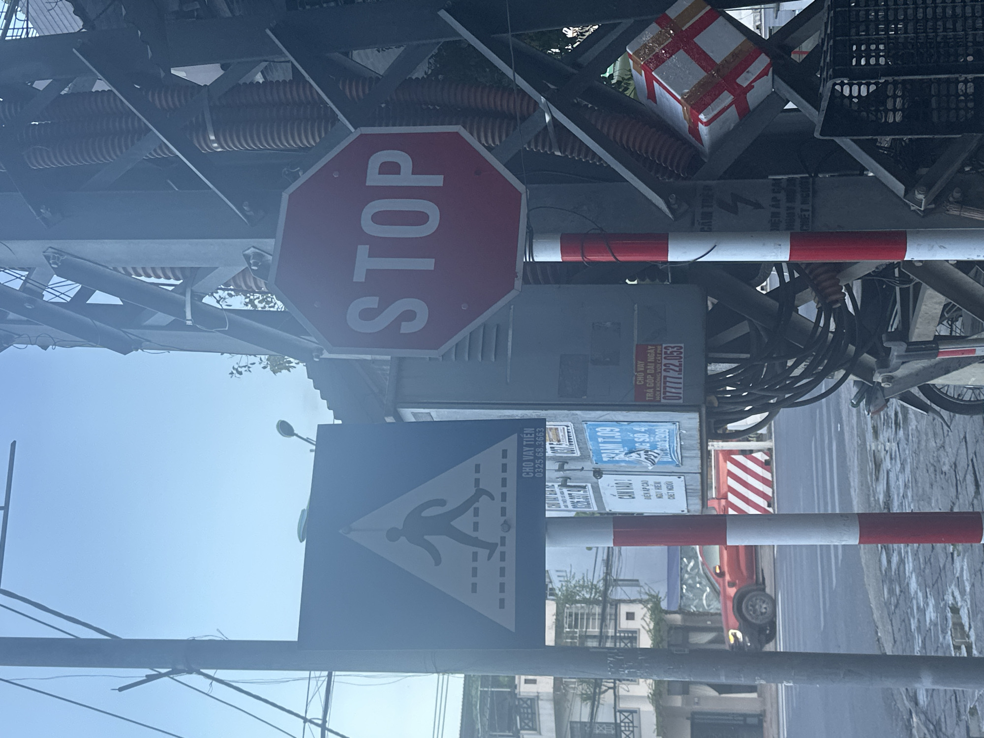 The image captures a street scene in Nam Nha Trang Ward, Vietnam. A prominent red octagonal stop sign is mounted on a metal pole, facing the viewer. To the left of the stop sign, a blue square sign with a white triangular pictogram of a pedestrian crossing is visible. The pedestrian sign is situated on a separate pole. Both poles are striped with alternating red and white sections at their bases. In the background, a red pickup truck is parked at a crosswalk. Various electrical equipment, including wires, conduits, and insulators, are attached to a large metal structure behind the stop sign. A white box wrapped with red tape is affixed to this metal structure. The ground appears wet, suggesting recent rain. Buildings line the street in the distance.
