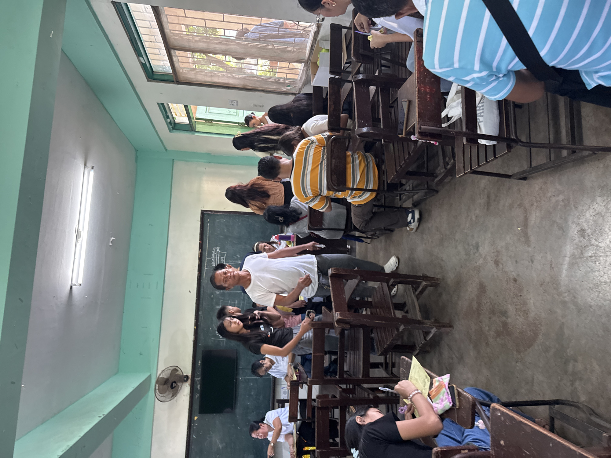 The image depicts an indoor scene, likely a classroom or multi-purpose room, in Davao City, Philippines, during daylight hours. The room features light mint green and white painted walls, a grey concrete floor, and numerous wooden desks and chairs. Natural light illuminates the space through louvered windows that show hints of foliage and buildings outside. Long fluorescent lights are mounted on the ceiling, and a wall-mounted electric fan is visible on one of the side walls.
Numerous individuals are present, suggesting a class, meeting, or assembly. In the central foreground, a man in a white t-shirt and grey pants stands, facing towards the right, appearing to be speaking or interacting with others. Near him, a woman with long dark hair stands, looking at her mobile phone. Most other people are seated at the desks; some are observing the front of the room, while others are focused on their phones or papers.
In the upper right corner, a person wearing a blue and white striped polo shirt is seen from behind. In the lower right corner, another individual in a black shirt is visible, looking at yellow papers on their desk. A white tote bag, partially visible between desks, has the word "PARK" printed on it. A dark blackboard or whiteboard in the background shows some very faint, mostly illegible writing, including what appears to be "Malabuyan". The atmosphere suggests an active and engaged gathering.