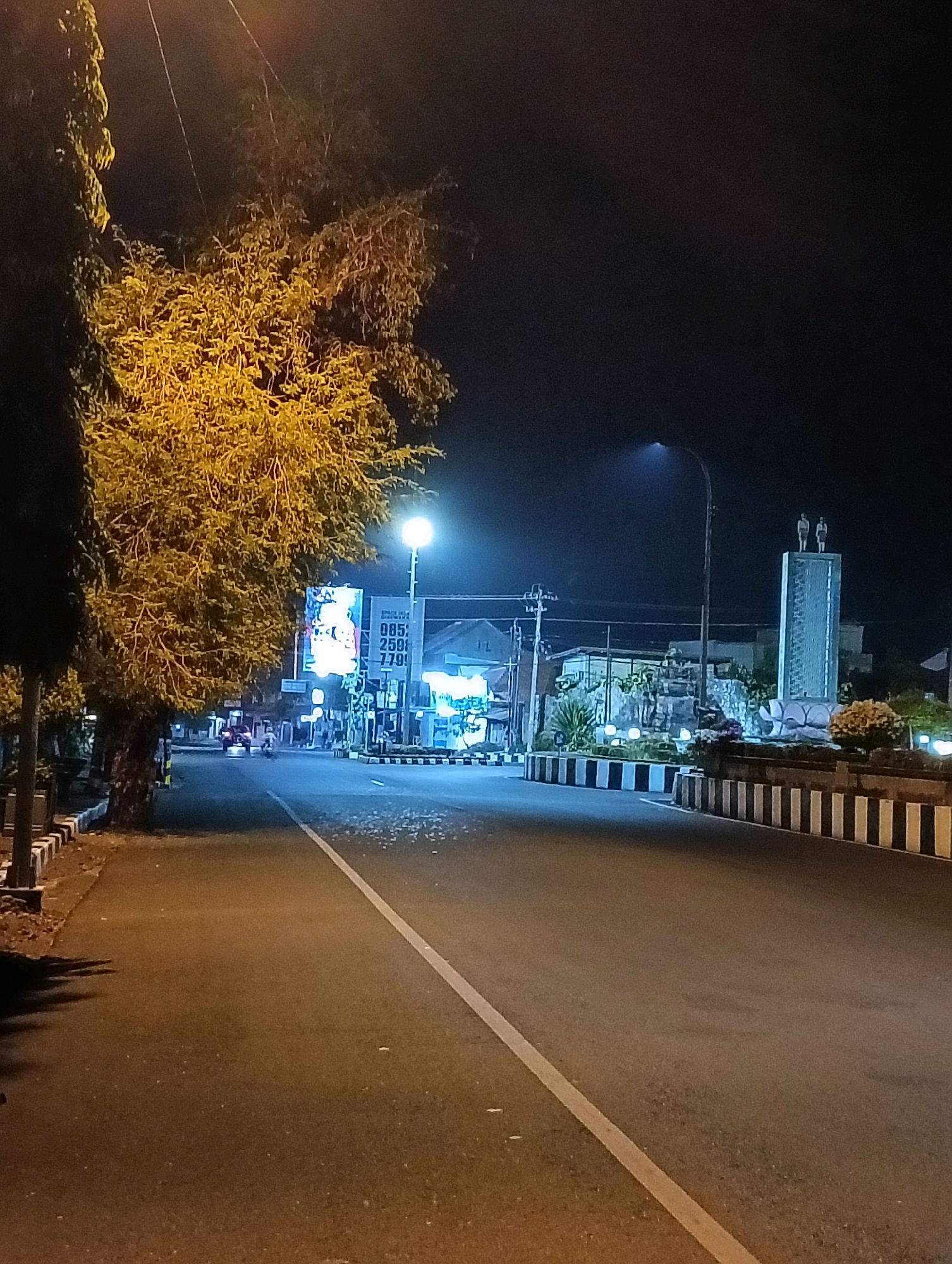 The image depicts a night scene on a street in Purworejo, Indonesia. A road with a white dividing line occupies the foreground. To the left, a tree with yellow-lit foliage stands beside a sidewalk with white and black striped borders. Further down the street, a billboard with Indonesian text and an advertisement is visible, illuminated by artificial light. On the right side of the road, a monument featuring two statues atop a column is prominently displayed, surrounded by decorative plants and lighting. The background shows buildings and streetlights, contributing to the urban night ambiance.