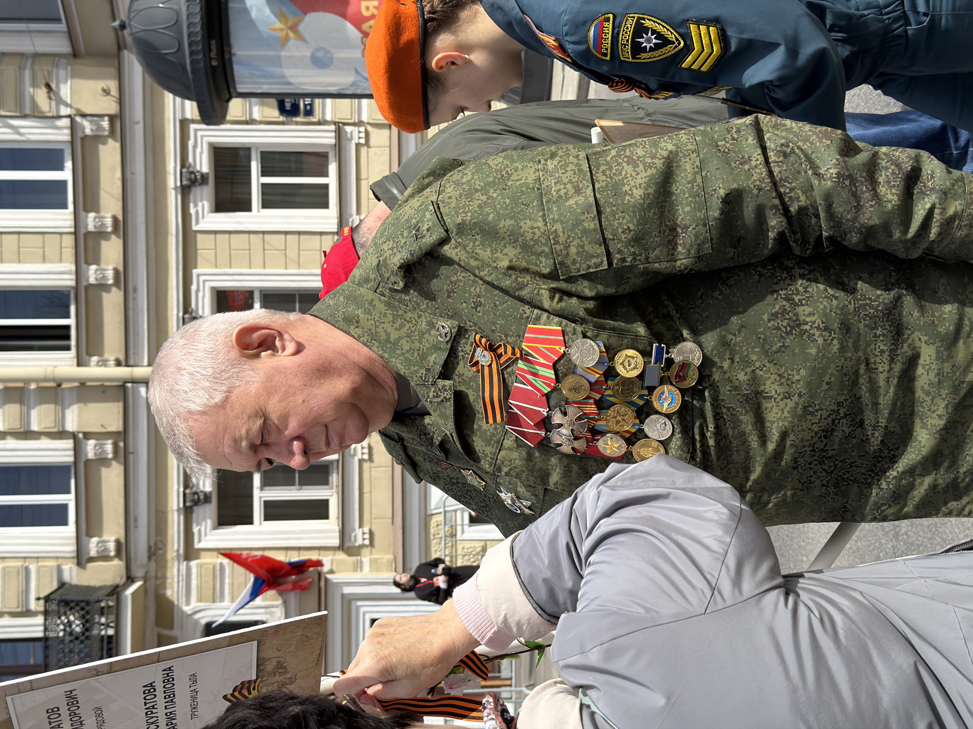 An elderly man in a camouflage uniform adorned with numerous medals stands in front of a building in Saint Petersburg, Russia. He is looking down at a sign being held by someone in front of him, while a woman with an orange beret stands in the background.