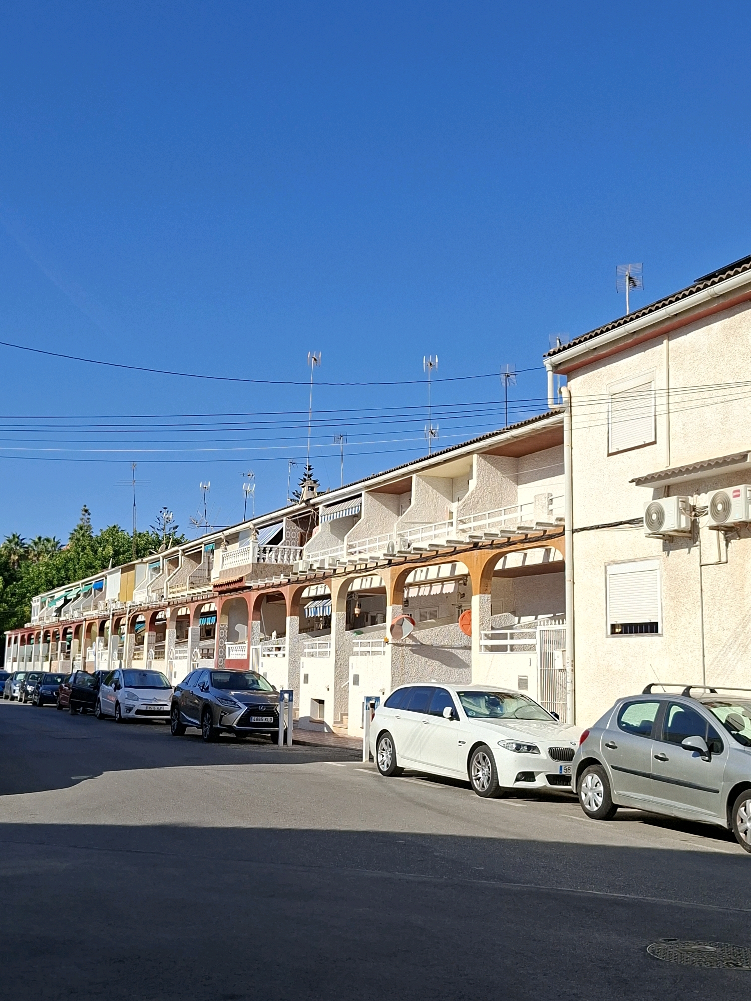 The image depicts a street scene in Torrevieja, Spain, characterized by a row of terraced houses and parked cars under a clear blue sky. The houses feature a light-colored stucco exterior with arched doorways on the ground floor, balconies with white railings on the upper floors, and striped awnings extended from some windows. Several television antennas are visible on the rooftops, along with air conditioning units attached to the facade of one building.
Parked along the street are several cars, including a silver Lexus RX with license plate "4665 KLD", a white BMW wagon, and a silver Peugeot. The road surface is asphalt, and a shadow of a vehicle falls across the foreground. Lush green trees are visible in the background, suggesting a residential area. The overall impression is one of a sunny, quiet residential street.