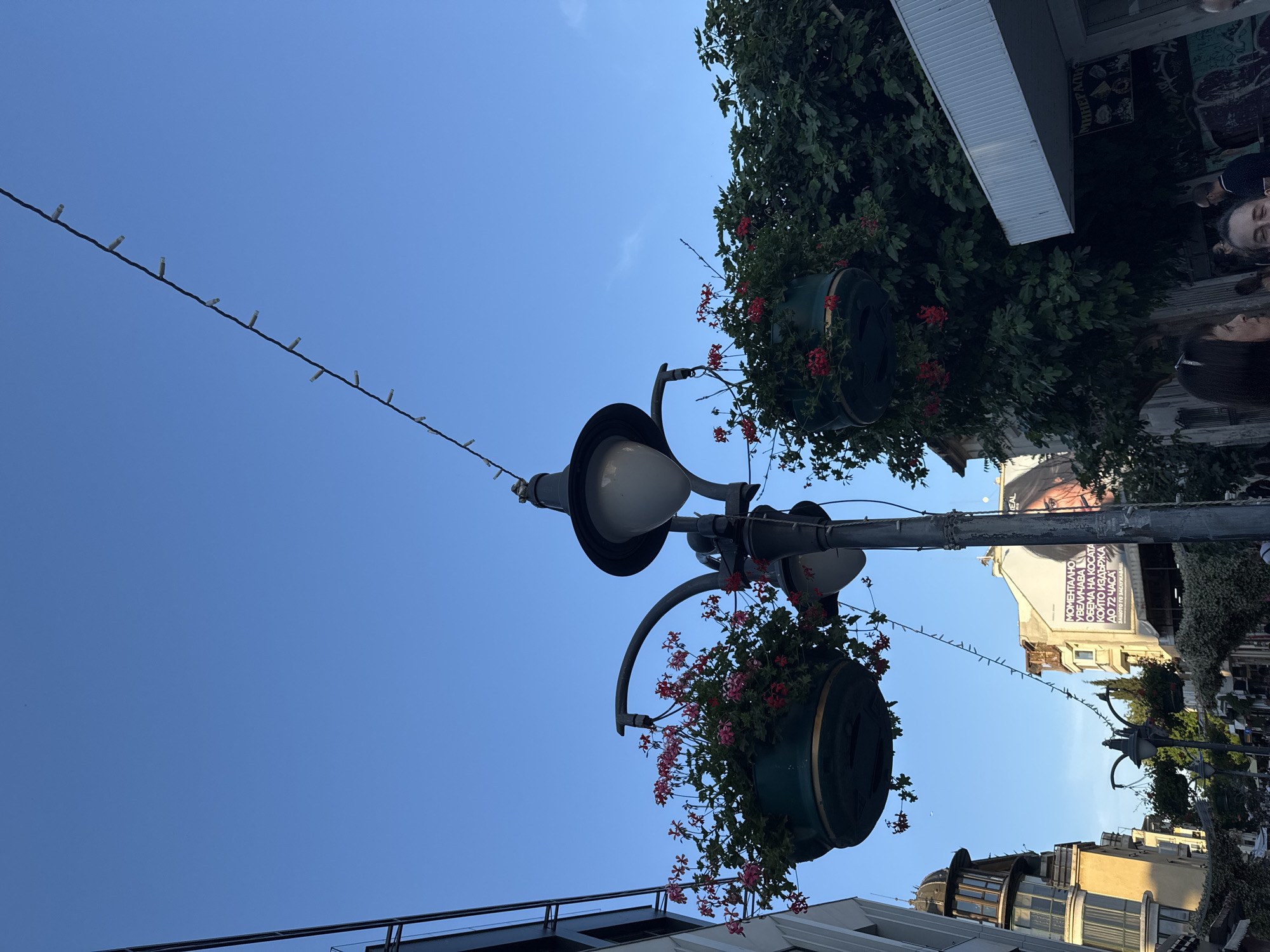 This is a street-level, medium-angle shot of the pedestrian zone in the city of Burgas, Bulgaria. The scene is illuminated by natural light under a clear blue sky. In the foreground, a lamppost with hanging flower baskets filled with red and pink flowers stands to the right of the frame. A string of small white lights hangs diagonally across the sky. To the left, a modern building with a dark facade and a prominent window structure is visible. Further down the street, buildings of varying architectural styles line the pedestrian walkway, with a large advertisement featuring a woman's face prominently displayed on one of the buildings. In the lower right corner, people are gathered, some partially visible, creating a sense of activity in the urban environment. The overall impression is a vibrant street scene in a European city.
