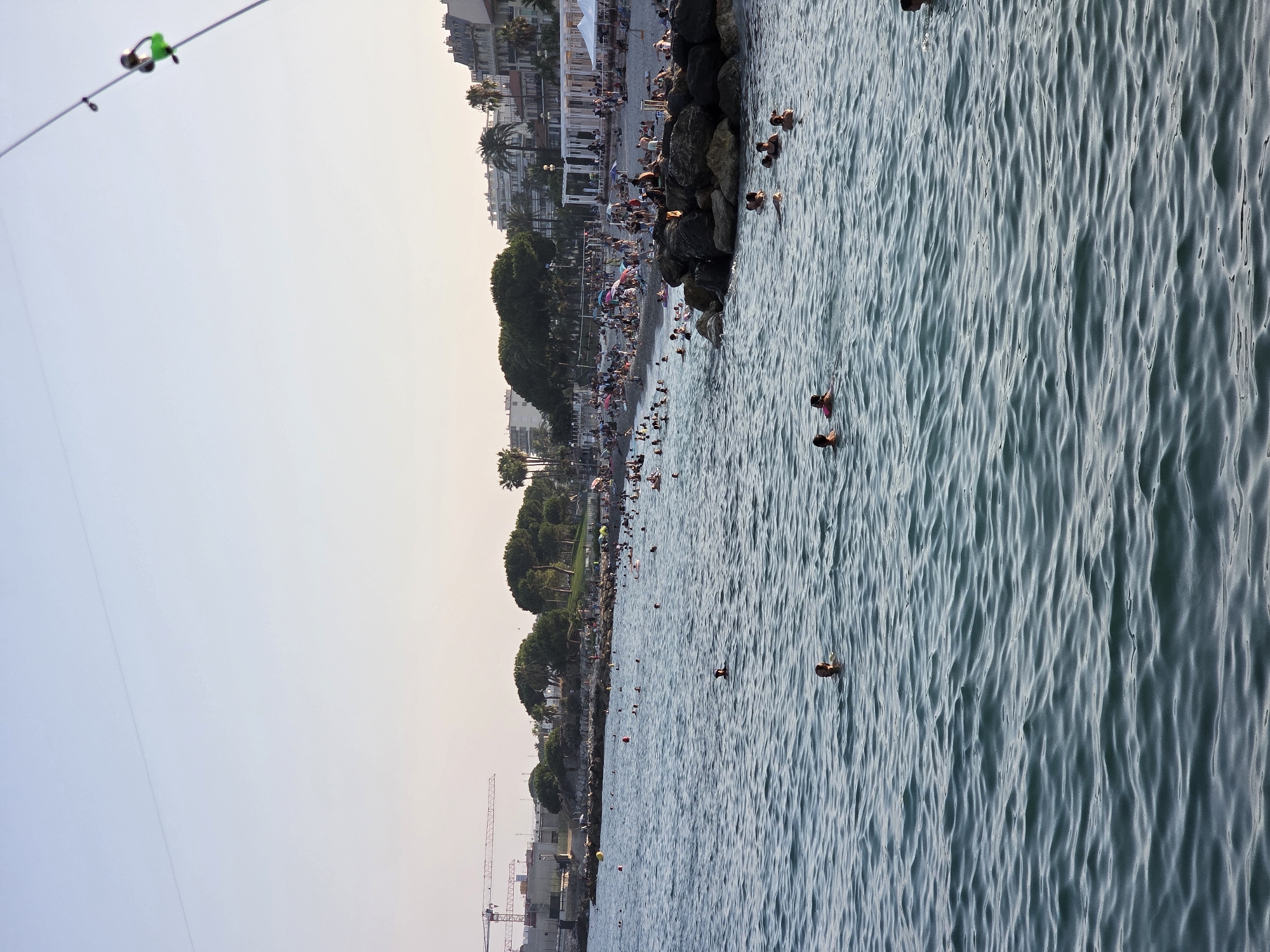 The image depicts a crowded beach and ocean scene in Nice, France, likely on the Promenade des Anglais, at the end of the day. The sky is a clear, pale blue, with a thin contrail visible. In the foreground, the turquoise ocean is rippled, with several people swimming. Further back, the pebble beach is packed with sunbathers under umbrellas, as well as people wading and swimming near the shore. Behind the beach, buildings line the coast, with palm trees scattered amongst them. Construction cranes are visible in the distance on the left, suggesting ongoing development. A fishing rod with a green bobber is prominently displayed in the upper right corner of the frame.