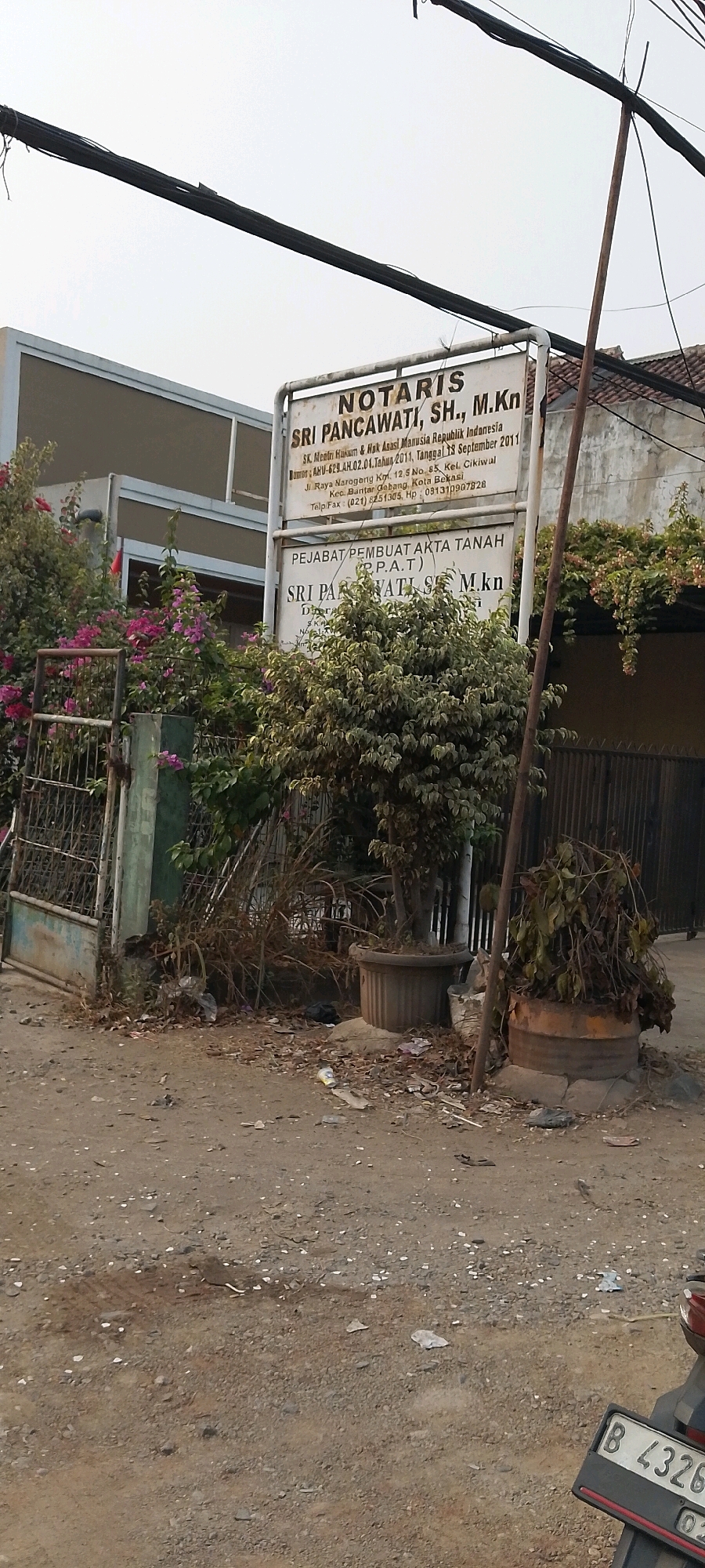 A medium-shot photograph captures a street scene in Bekasi, Indonesia, focusing on a notarial office. The sign for "NOTARIS SRI PANCAWATI, SH., M.Kn" is prominently displayed, with text detailing her legal qualifications and address at Jl. Raya Narogong Km. 12.5 No. 85, Kel. Cikiwul, Kec. Bantar Gebang, Kota Bekasi. Below this, another sign indicates "PEJABAT PEMBUAT AKTA TANAH (P.P.A.T.) SRI PANCAWATI, SH., M.Kn". The surrounding environment is somewhat cluttered, with dried leaves, trash, and two large potted plants situated near the building's entrance. A metal gate and fence are visible to the left, partially covered by flowering pink bougainvillea. Power lines are strung overhead, and a motorcycle license plate reading "B 4326" is visible in the lower right corner. The ground is unpaved and appears to be a dirt road or parking area. The lighting suggests it is daytime.