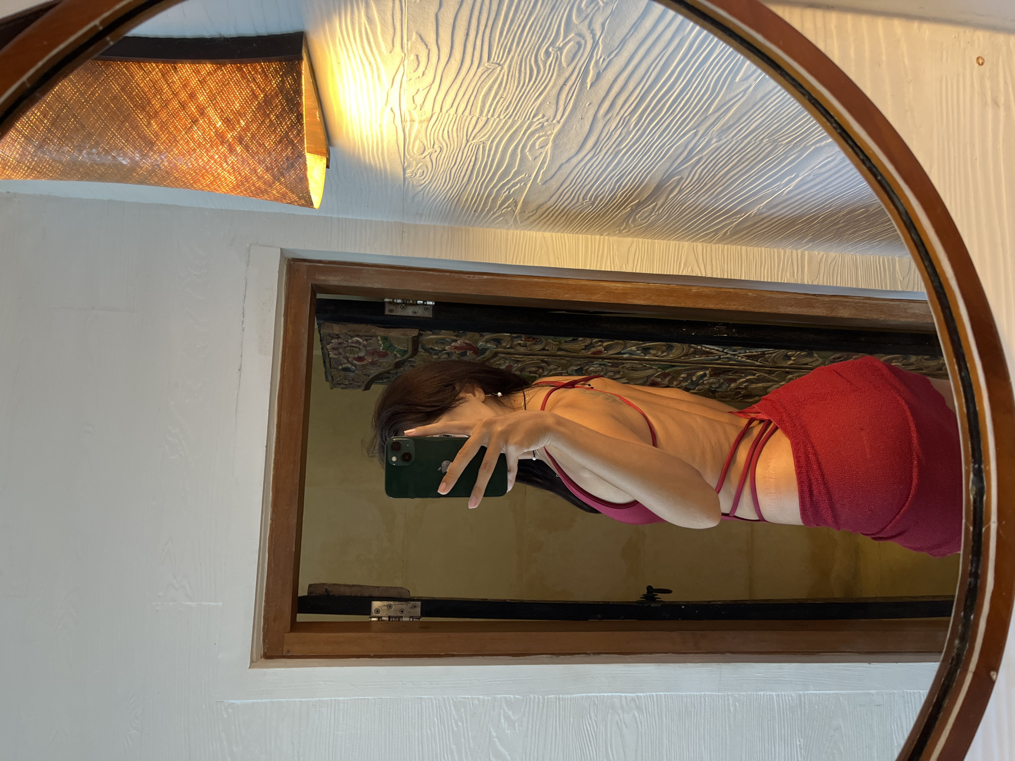 An indoor, vertically oriented mirror selfie captures a woman in a rustic-style room, located in Kuta Selatan, Indonesia. The central subject is a young woman, visible from the waist up in a round, wooden-framed mirror. She has dark hair and is wearing a dark red, backless garment with thin, crisscrossing straps on her lower back. She holds a dark green smartphone (likely an iPhone with a triple-camera setup) to take the photograph, obscuring part of her face.
The room features white, textured walls and a white ceiling with a distinct, wavy, wood-grain-like pattern. A woven, possibly rattan or bamboo, wall sconce with a warm light illuminates the upper left area of the reflection, casting light onto the textured ceiling. Inside the main mirror's reflection, behind the woman, is a wooden-framed opening or window, and beyond it, a glimpse of an intricately patterned, possibly carved or textile, decorative object is visible. The warm artificial lighting suggests it might be evening or an interior scene. No visible text is present in the image.