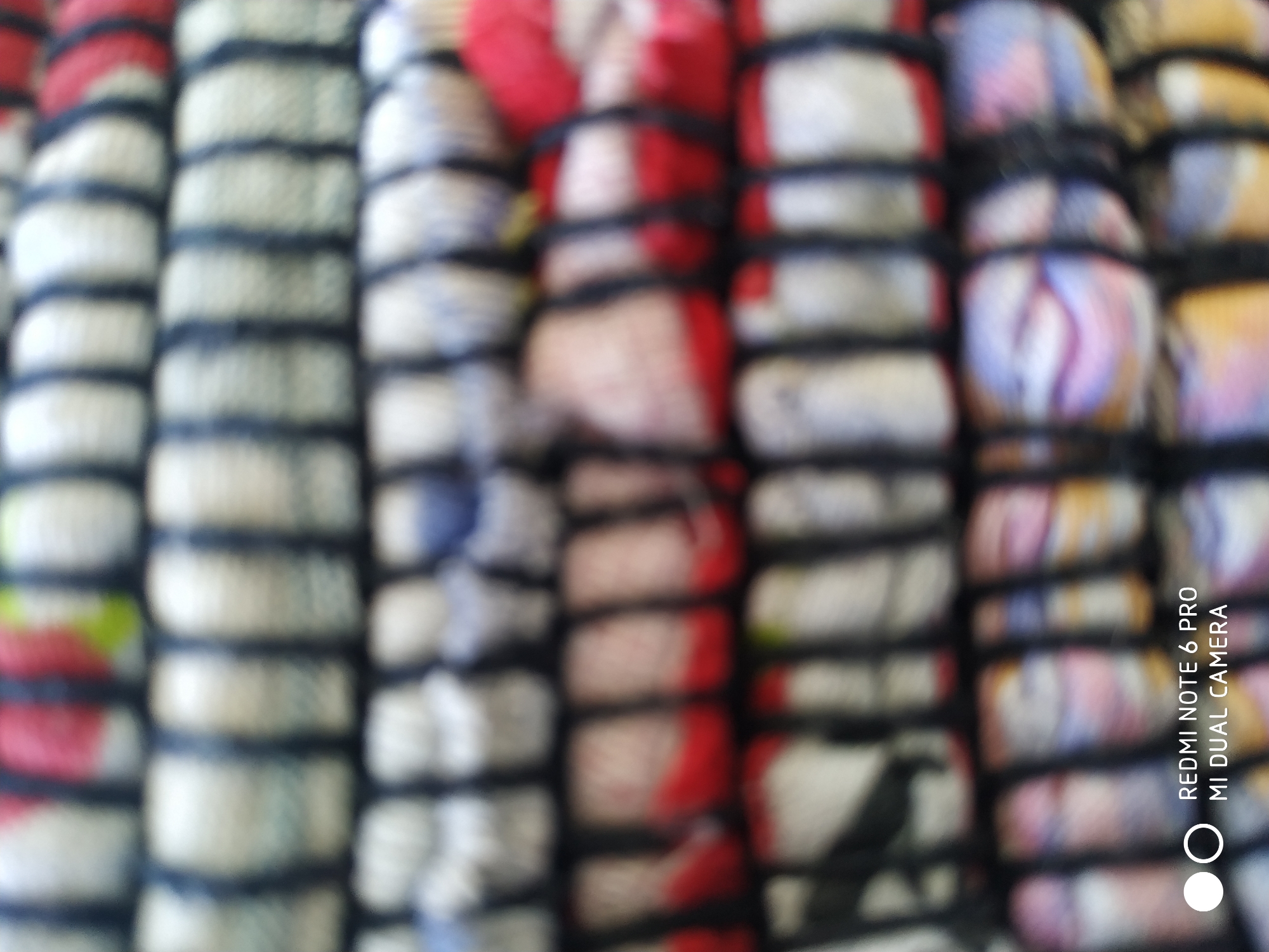 The image is a blurry, close-up shot of a stack of colorful woven items, possibly rugs or fabric rolls. The items are layered horizontally, with dark vertical bands separating each layer. The colors visible include red, white, blue, green, and yellow, suggesting a floral or patterned design. The texture appears to be rough and fibrous. There are no people or discernible environmental features. The bottom left corner of the image contains text that reads "REDMI NOTE 6 PRO MI DUAL CAMERA," indicating it is a photograph taken with that device. The image does not provide any information about the location in Fryazino, Russia, or any other geographical context.