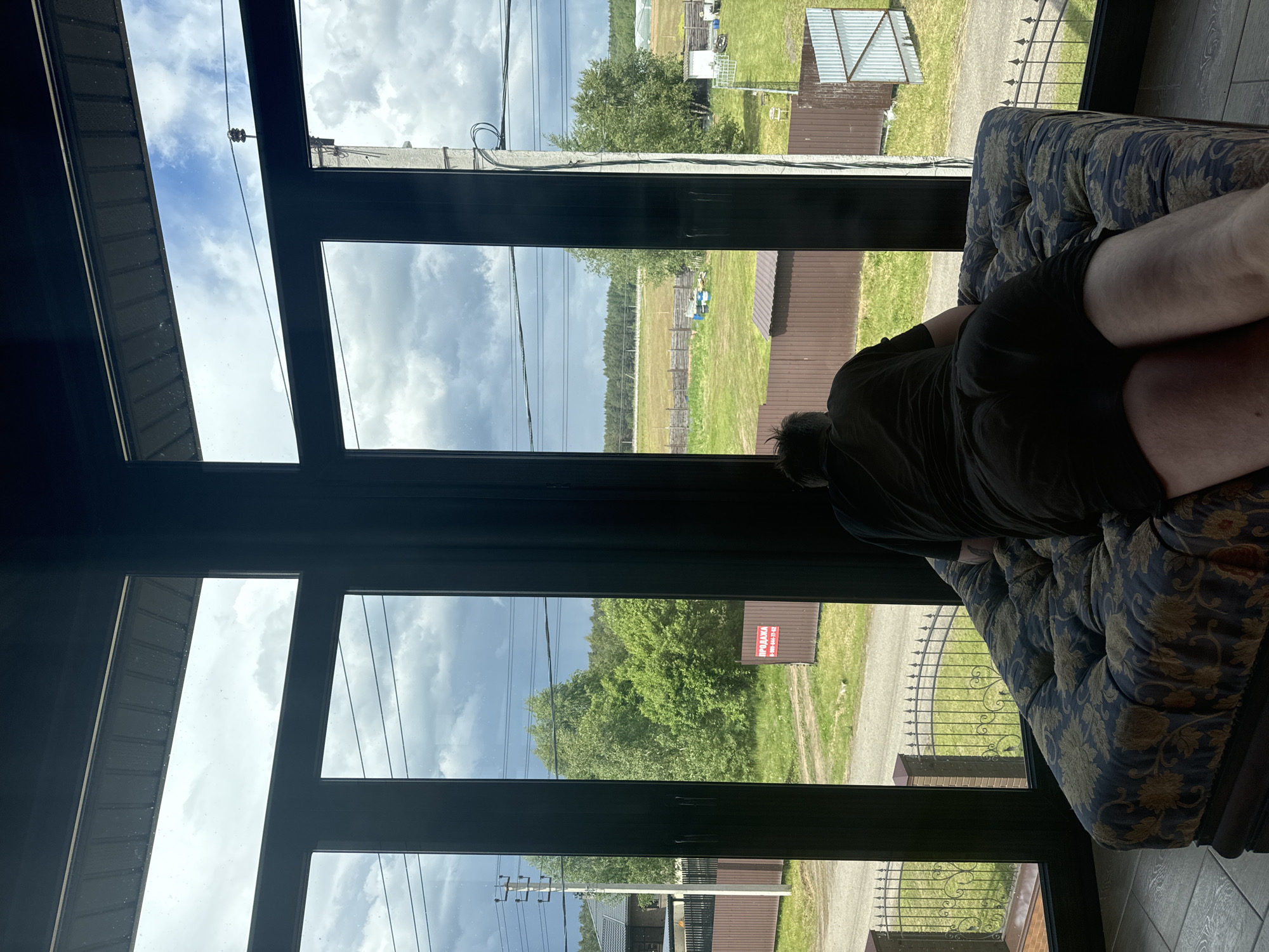 A person in dark clothing lies on a floral-patterned couch indoors, gazing through a multi-paned glass window. The view outside includes a rural landscape with green grass, trees, fences, and power lines. In the distance, buildings are visible. A sign in Cyrillic script is also seen. Based on the location provided (Макарово, Russia), the scene suggests a rural or suburban setting on the outskirts of the city.