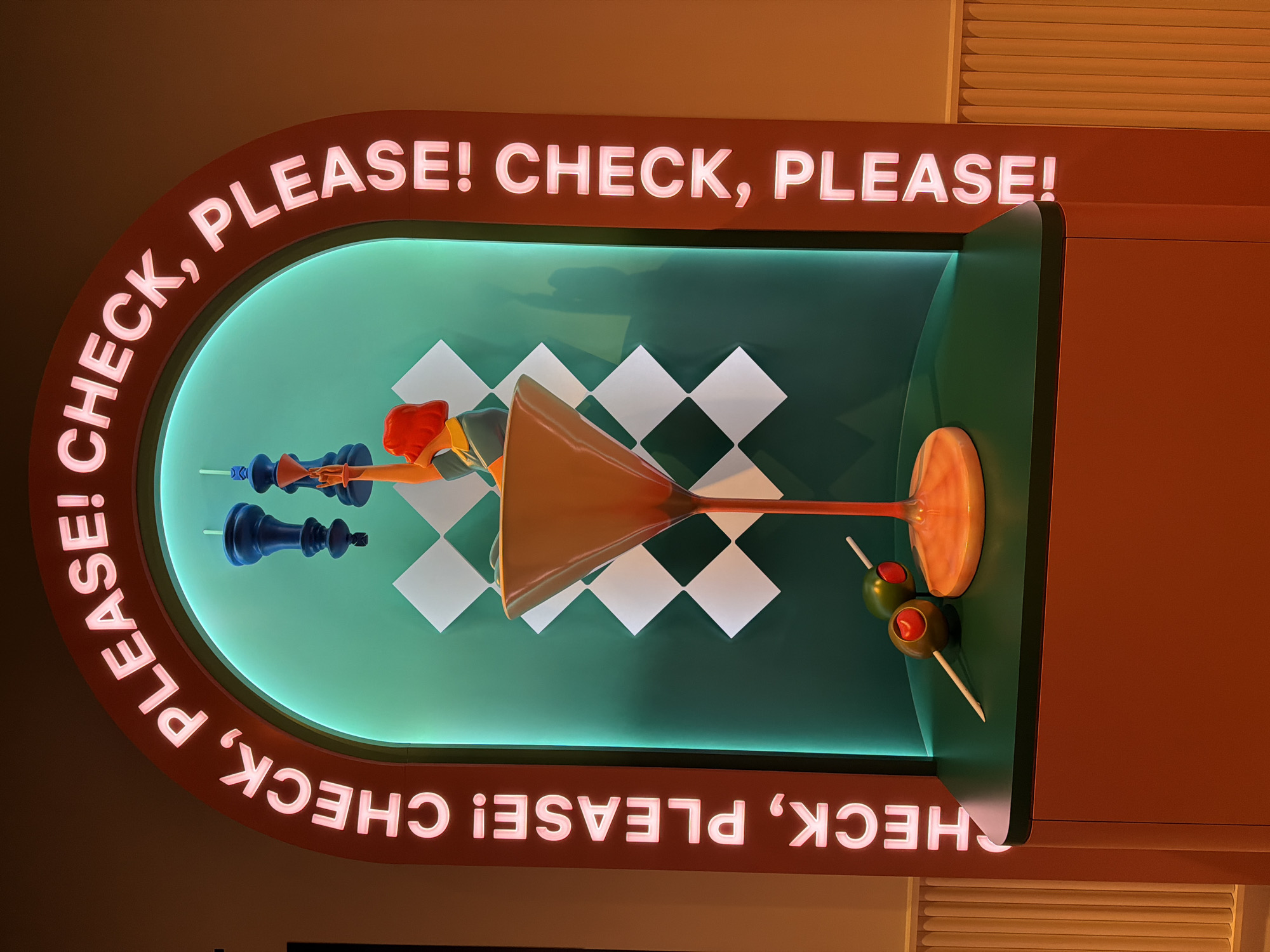 The image depicts a neon sign with the words "PLEASE! CHECK, PLEASE!" framing a display case. Inside the case, there is a stylized arrangement featuring chess pieces, a martini glass, and olives against a backdrop of white and black squares. The scene is located in the city of Dallas, United States of America.