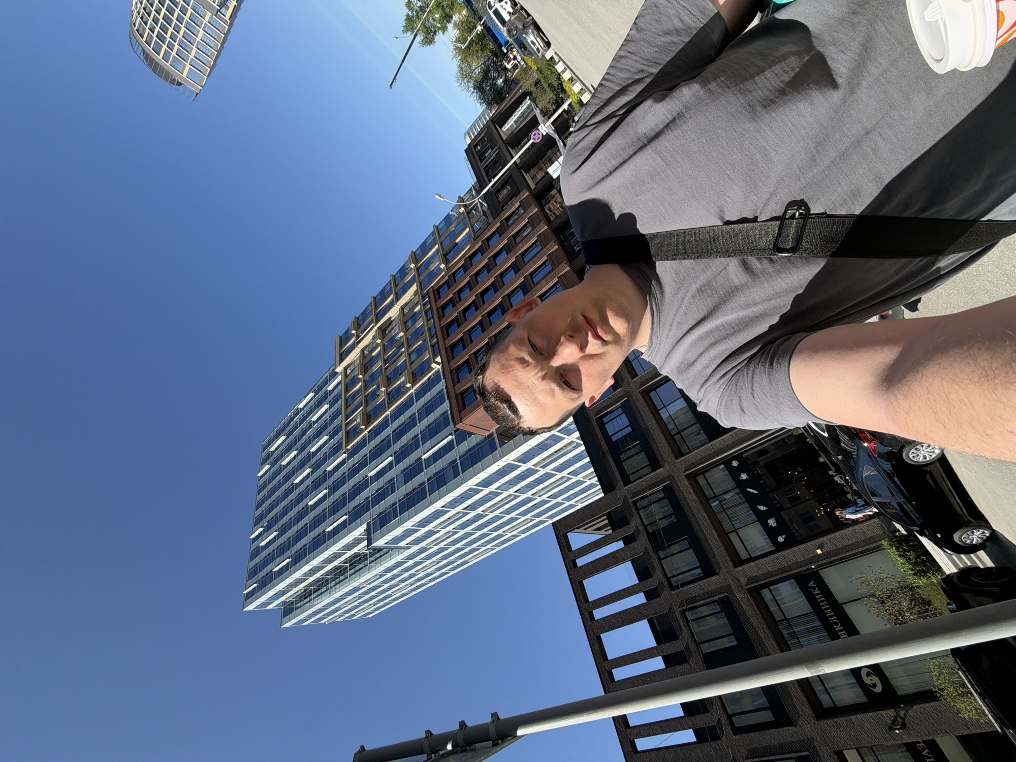 A first-person perspective photograph shows a man wearing a gray shirt and a black crossbody bag, standing outdoors on a sunny day. The background features a cityscape in Yekaterinburg, Russia, including several modern buildings: a skyscraper with a white and blue grid-like facade, a brick-colored high-rise building with many windows, and a darker building with a structured grid pattern. Street signs, light poles, trees, and parked vehicles are also visible.