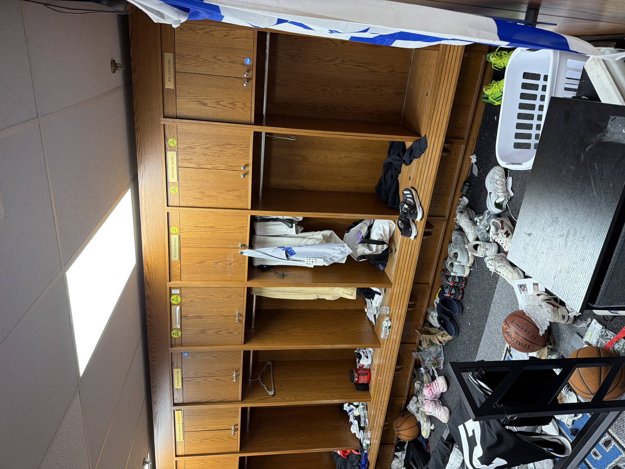 The image captures an indoor locker room, primarily focusing on a long bank of wooden lockers. The lockers are arranged in vertical columns, each typically featuring a small closed cabinet at the top and an open cubby shelf below. Many of the cubbies contain clothing, towels, and various personal items, while some are empty.
The floor beneath the lockers and extending into the foreground is carpeted and appears quite cluttered. Numerous pairs of athletic shoes, including sneakers and slides, are scattered across the floor, along with plastic water bottles, plastic bags, and other debris. Several basketballs are visible, one prominently displaying the "SPALDING" brand. A white, rectangular plastic laundry basket sits near the right side of the frame. Above, a drop ceiling with white acoustic tiles and bright fluorescent light fixtures is visible. The overall scene suggests the locker room is actively used, possibly after a sports event or practice.
Visible text on locker labels includes "#33 Johny Dan," "#34 Nate Jacobs," and "#13 Roy Itcovichi." The brand name "SPALDING" is legible on a basketball. The OCR also detected "TE-TRAINER," though its specific placement in the image is not clearly discernible.