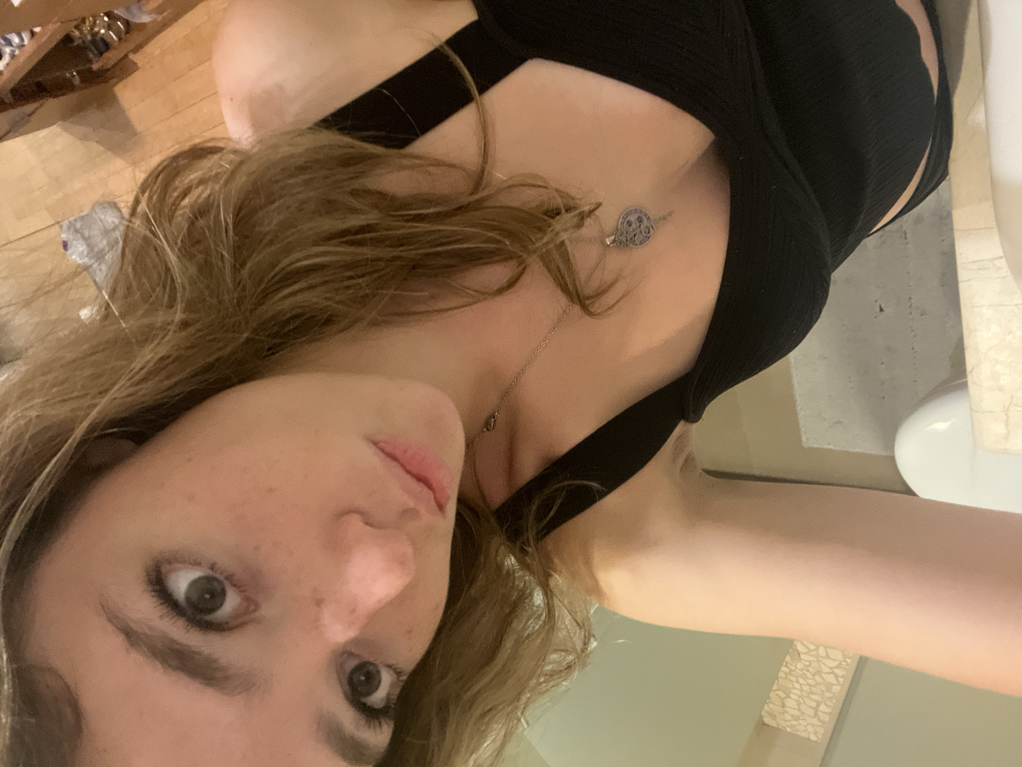 A close-up, angled selfie of a light-skinned female with freckles and brown hair, wearing a black tank top and a delicate silver necklace with a round pendant. The background includes a wooden floor, a partially visible cabinet or shelf, and a white sink with a light-colored counter. No landmarks visible to identify San Bernardino Tlaxcalancingo, Mexico.