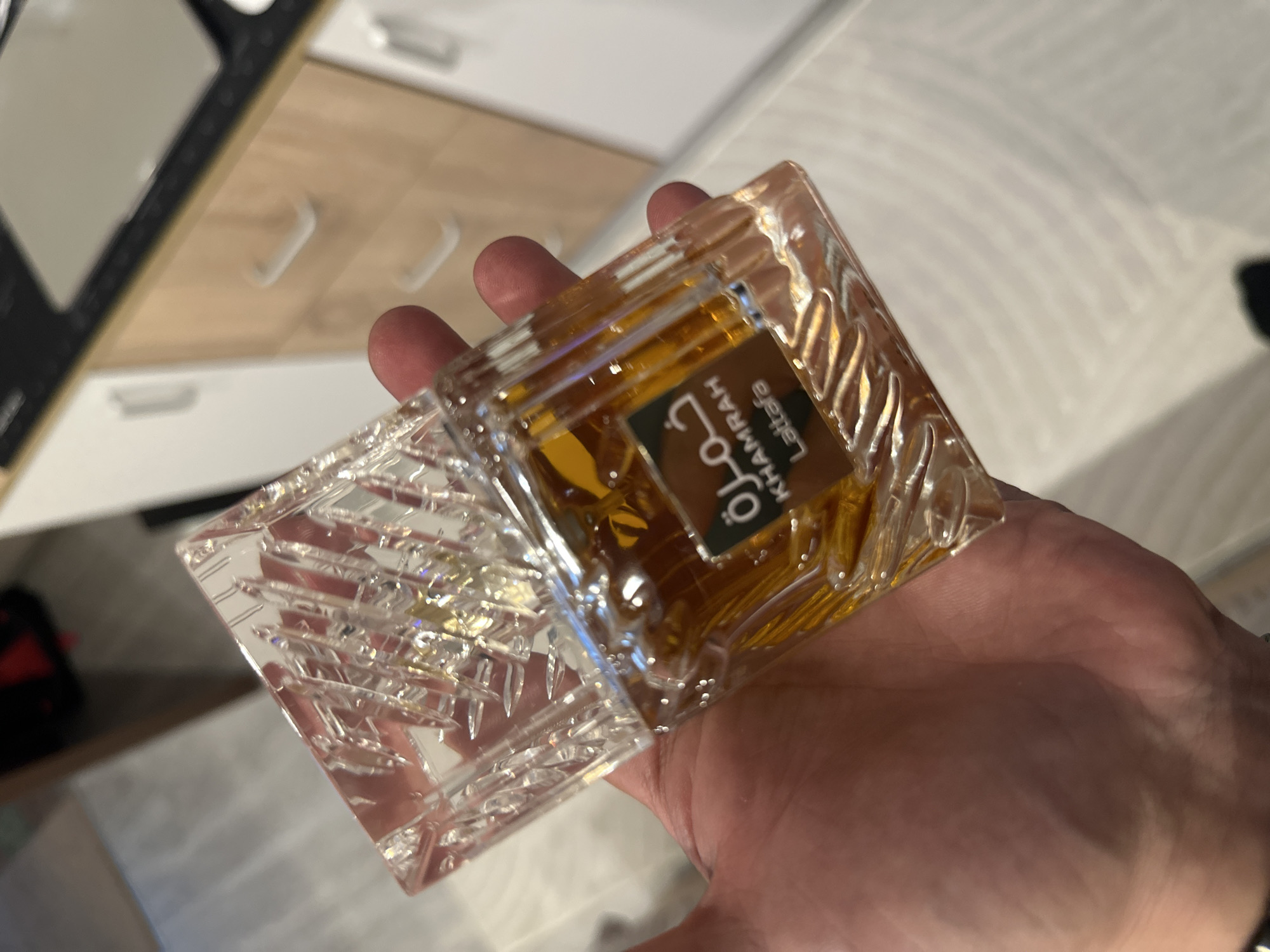 A hand holds a bottle of Lattafa Khamrah perfume, its cut-crystal design catching the light. The bottle is partially open, with the cap resting near the base. The background suggests an interior setting with a kitchen area visible in the periphery, including cabinets with drawer pulls.