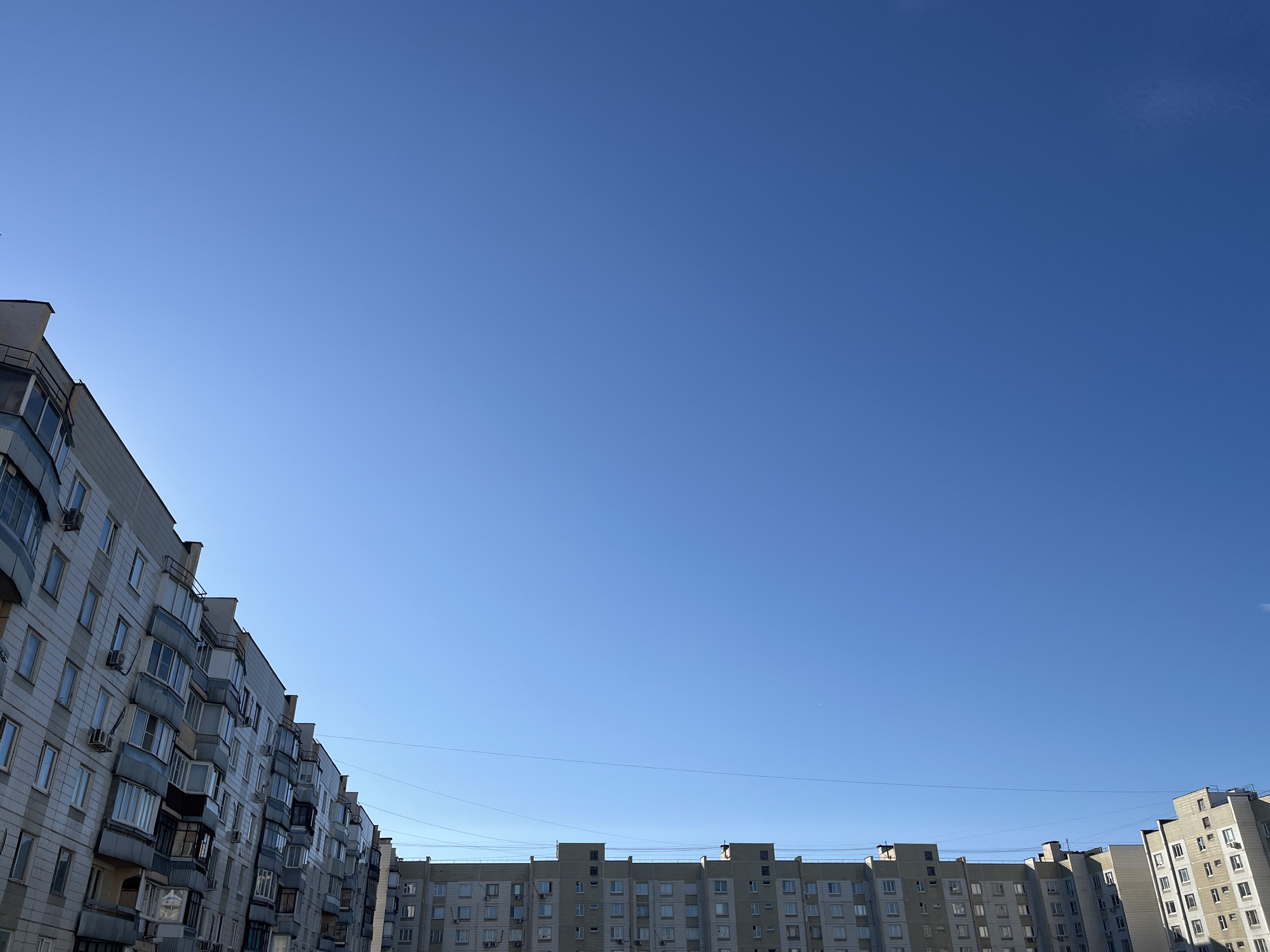 This is an outdoor, eye-level, medium shot of a clear blue sky with two apartment buildings visible on the left and right sides of the frame. The apartment buildings are of medium height, with multiple stories and numerous windows. The building on the left appears to be an older, gray concrete structure with a facade that has some texture. The building on the right is lighter in color, possibly concrete or stucco, and appears to be of a similar architectural style with multiple stories and windows. Both buildings are residential, and there are air conditioning units visible on many of the windows. There are also balconies with railings on the building on the left. Thin black wires, likely power or communication lines, are strung between the buildings. The sky is a uniform, bright blue with no clouds. The lighting suggests it is daytime, likely morning or afternoon, with the sun to the left of the frame, casting some shadows on the left building. The scene is devoid of people or any visible activity. The location is likely a residential area in Moscow, Russia, given the architectural style of the apartment buildings, which are characteristic of Soviet-era housing blocks.