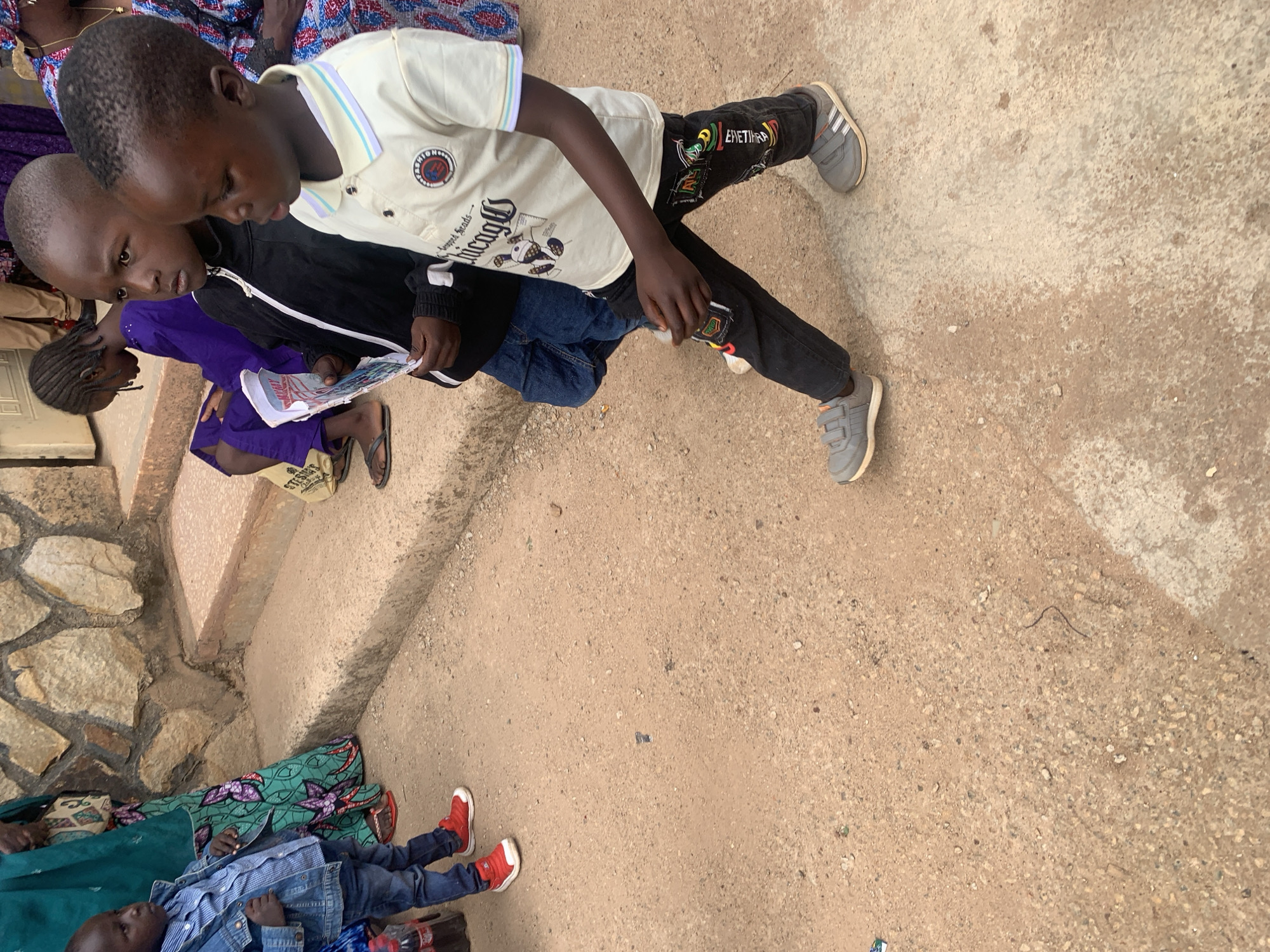 **Scene:** A partly-rotated shot taken outdoors. The primary subjects are two young boys, one wearing a cream shirt with a circular logo and "Chicago" script, and the other in a black jacket and jeans. They are focused on reading a newspaper or magazine together.
**People:** The boys are positioned on a rough, uneven terrain with scattered debris. Other people are present but partially cropped; an individual in purple is partially visible behind the boys. The lower portion of the image shows a child lying down in a blue denim jacket and red sneakers, adjacent to an adult wearing a green and floral-patterned garment.
**Environment:** The ground appears to be a mix of dirt and concrete, with portions of a wall and steps visible in the background.
**Location:** The picture appears to have been taken in Jos, Nigeria.