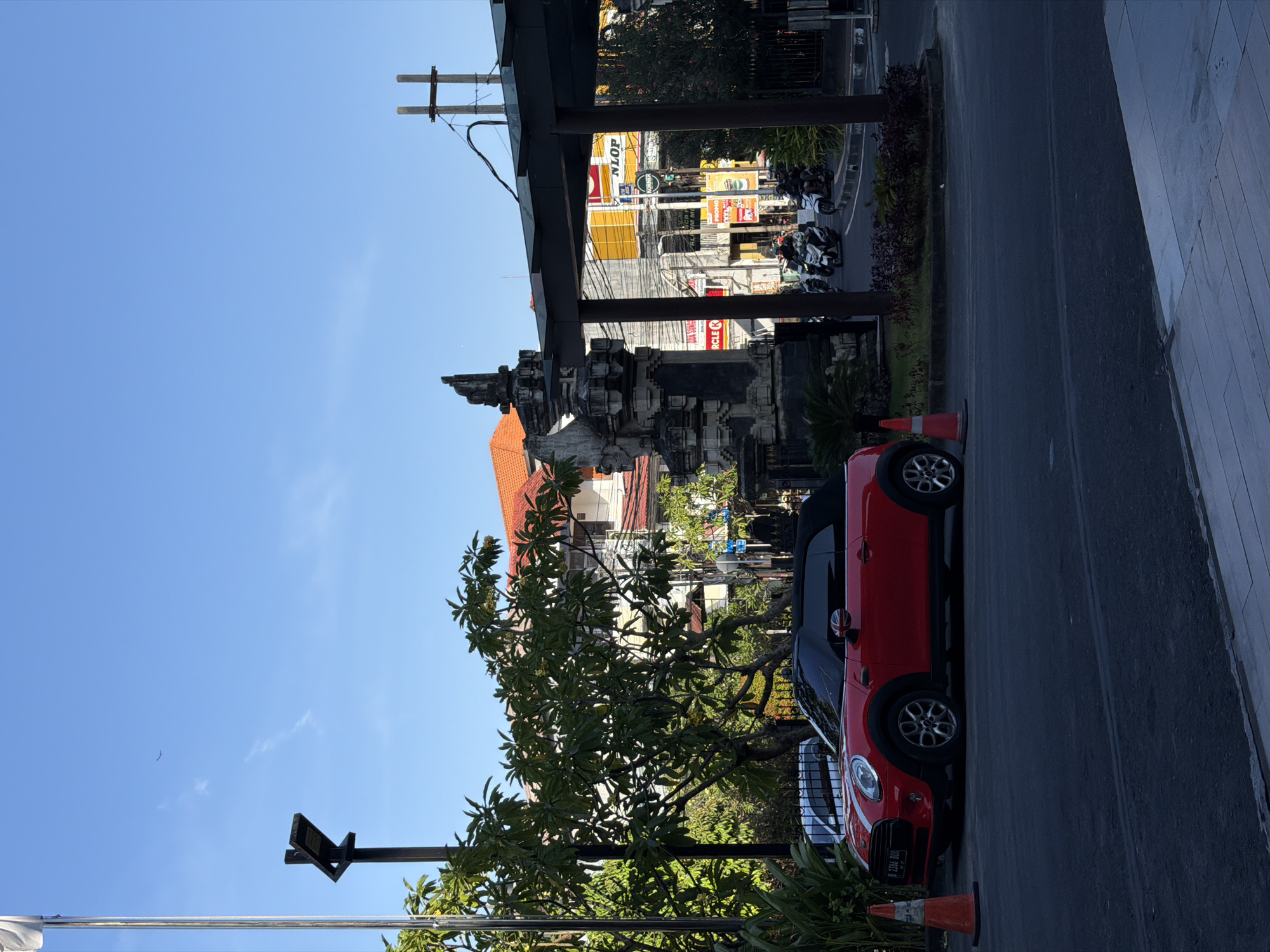 An eye-level, medium shot captures a red Mini Cooper convertible parked roadside in Denpasar, Indonesia. The car, positioned parallel to the viewer, has its top down and faces left. An orange traffic cone is placed in front of the vehicle, and another cone is visible behind it. Behind the Mini Cooper, a white car is parked.
The backdrop features a street scene with lush green trees and traditional Balinese architecture. A prominent stone archway, characteristic of Balinese temples, stands to the left of the frame. Beyond the archway, buildings with red-tiled roofs and multiple stories are visible. The street is active with several people riding motorcycles.
Signs for businesses like "NLOP," "KORNER," and "CIRCLE K" are displayed on buildings in the background. A utility pole with wires crisscrosses the scene. The sky is a clear blue, with a single bird soaring in the distance and a wispy cloud. The overall impression is a sunny day in a bustling Indonesian city, blending modern vehicles with traditional cultural elements.