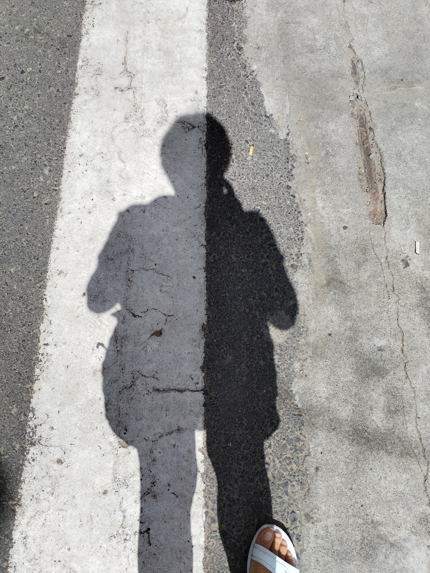 The image provides an overhead view of a person's shadow cast onto a pedestrian crossing or marked pathway. The shadow is bisected vertically by a prominent, worn white line painted on the ground. The left side of the shadow falls on a dark, textured asphalt surface, while the right side is cast onto lighter grey concrete that shows cracks and some small debris. Consequently, the shadow's left half appears lighter against the white stripe, and its right half appears darker on the concrete.
The lower right corner of the frame reveals the person's right foot, adorned with a white sandal, partially visible on the concrete. The sharp, well-defined nature of the shadow indicates bright sunlight, suggesting the photo was taken on a clear, sunny day, likely around midday or early afternoon. The cracked pavement and faded markings imply an outdoor urban or suburban setting within Bac Nha Trang Ward, Vietnam. The scene captures a momentary presence on a marked path, possibly during a walk. There is no visible text in the media file.