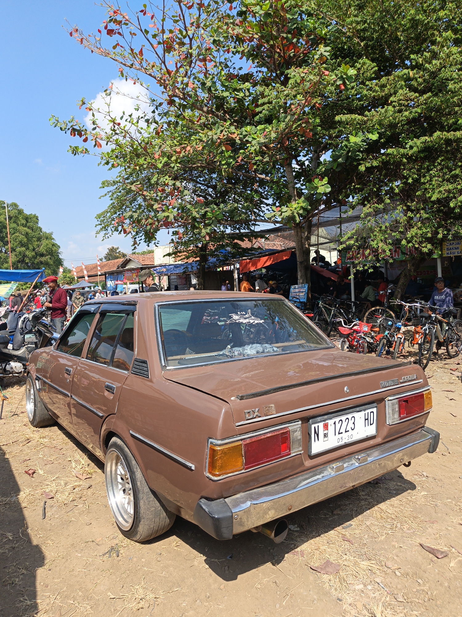 In Jatinom, Indonesia, under a bright blue sky with scattered clouds, a brown 1980s Toyota Corolla DX sedan is parked on a dirt ground littered with dry leaves and grass. The car, viewed from the rear-right quarter, has a chrome bumper, aftermarket rims, and a license plate reading "N 1223 HD". Behind the car, a vibrant green tree with some reddish leaves shades a bustling outdoor market scene. Several individuals are present, some standing near motorcycles, others seated under makeshift stalls adorned with orange and blue tarps. Numerous bicycles of various sizes and colors are parked nearby, suggesting a community gathering or local market. The overall atmosphere is one of a casual, everyday event in a rural Indonesian setting.