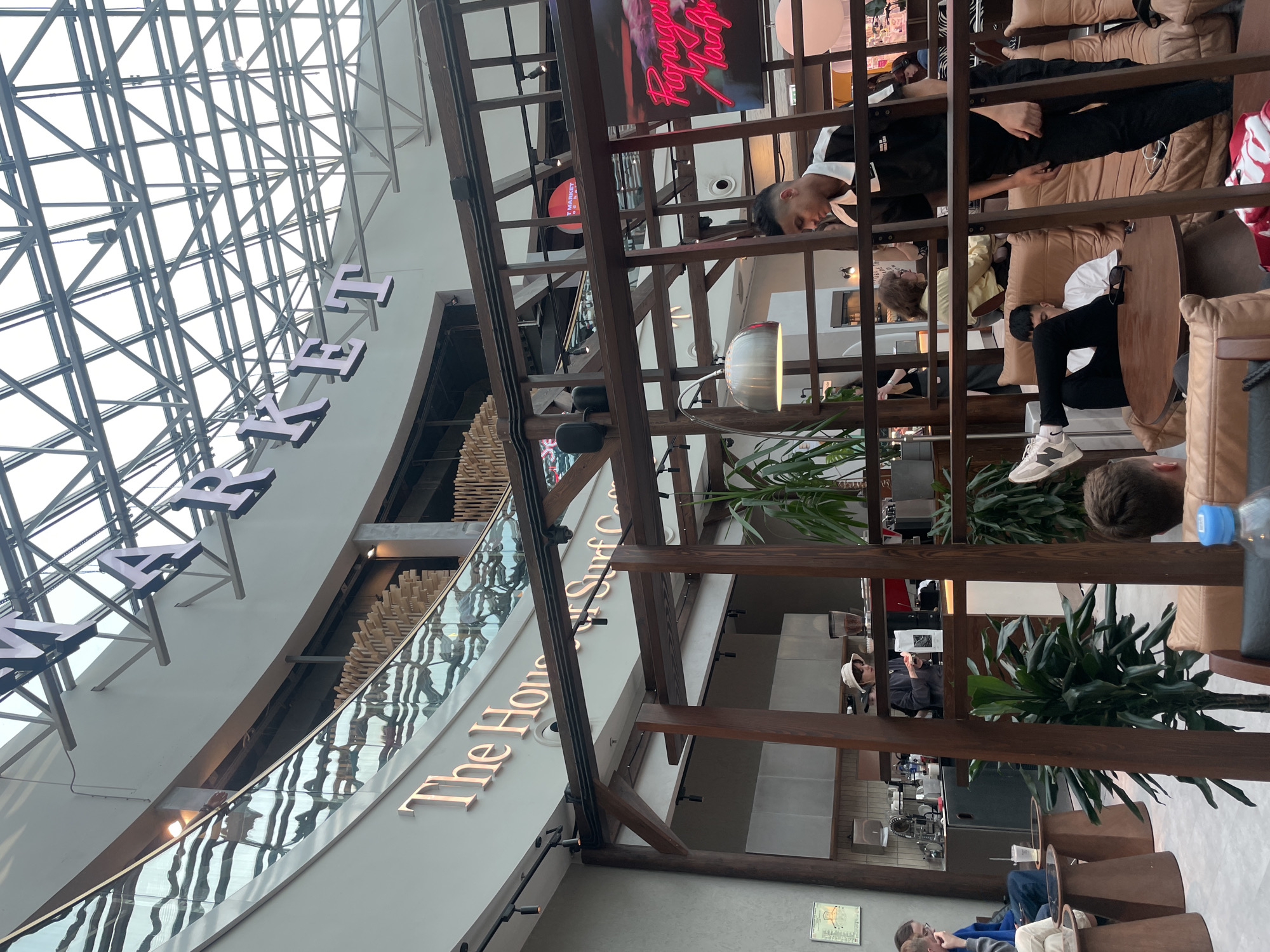 Here is a detailed description of the image:
**Environment**: The image shows the interior of a modern, well-lit space, likely a commercial area such as an airport or a shopping mall. A large glass ceiling with a grid-like structure provides ample natural light.
**Signage**: There is signage indicating "Market" and "The House of Surf & Cafe", suggesting the presence of stores and cafes.
**People**: Several people are present in the image. Some are sitting on sofas, engaging in activities like using phones or relaxing. Others are standing and walking around.
**Objects**: Various objects are visible, including sofas, tables, plants, wooden structures (possibly decorative or architectural), and a water bottle.
**Location**: Based on the signage and architectural style, the image is likely taken in Saint Petersburg, Russia, particularly inside the Pulkovo airport.
