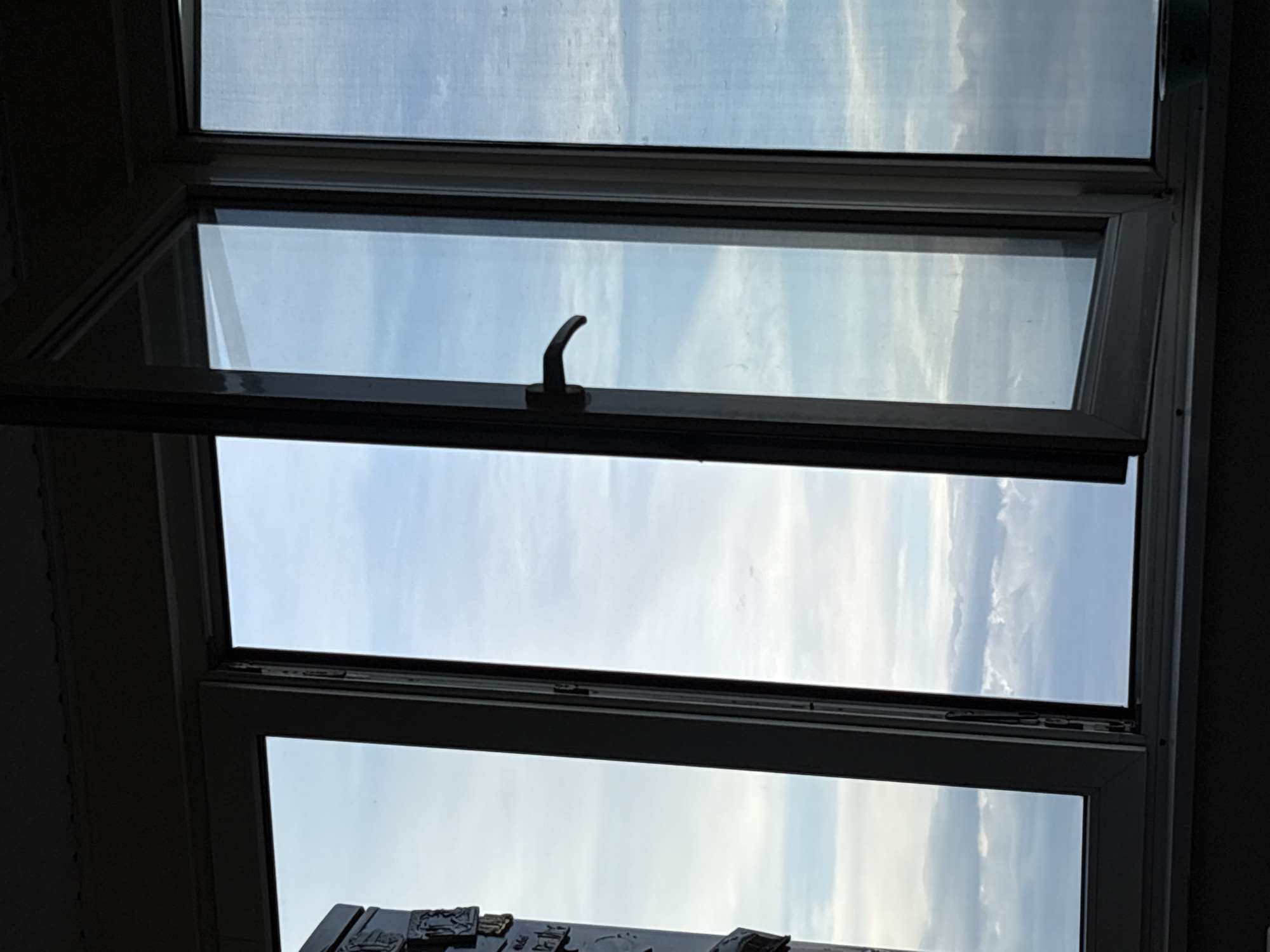 The image depicts a view through an open window, likely from an apartment or building in the city of Vratsa, Bulgaria. The frame of the window is visible, with one pane open at an angle, revealing a sky filled with white and blue clouds. The lower part of the window frame is darker, suggesting it is a sill or interior ledge. The upper part of the image is silhouetted, possibly indicating the ceiling or the exterior of the building. There are no people or specific landmarks visible that would further pinpoint the location within Vratsa. The scene is focused on the open window and the expansive cloudy sky.