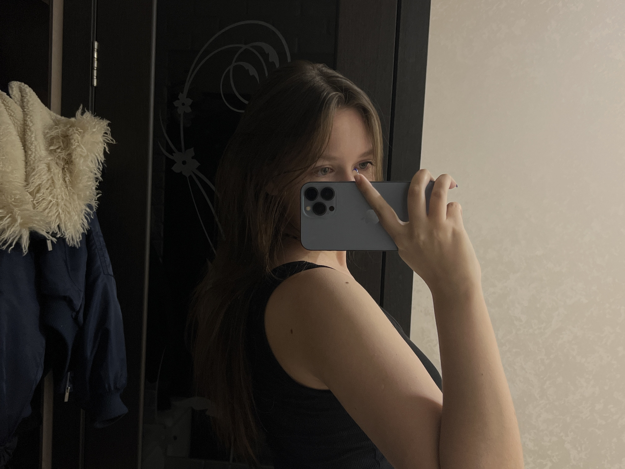 A woman is taking a selfie in what appears to be an indoor setting. She is holding a smartphone, obscuring part of her face with the device. She has light-colored eyes and long brown hair. She is wearing a black tank top. Behind her, there's a dark wardrobe or closet with some decorative patterns on the doors. A coat with a white fur collar hangs on the side of the wardrobe. The wall behind her is a light, textured beige. No specific details indicate the location is Chaikovsky, Russia.