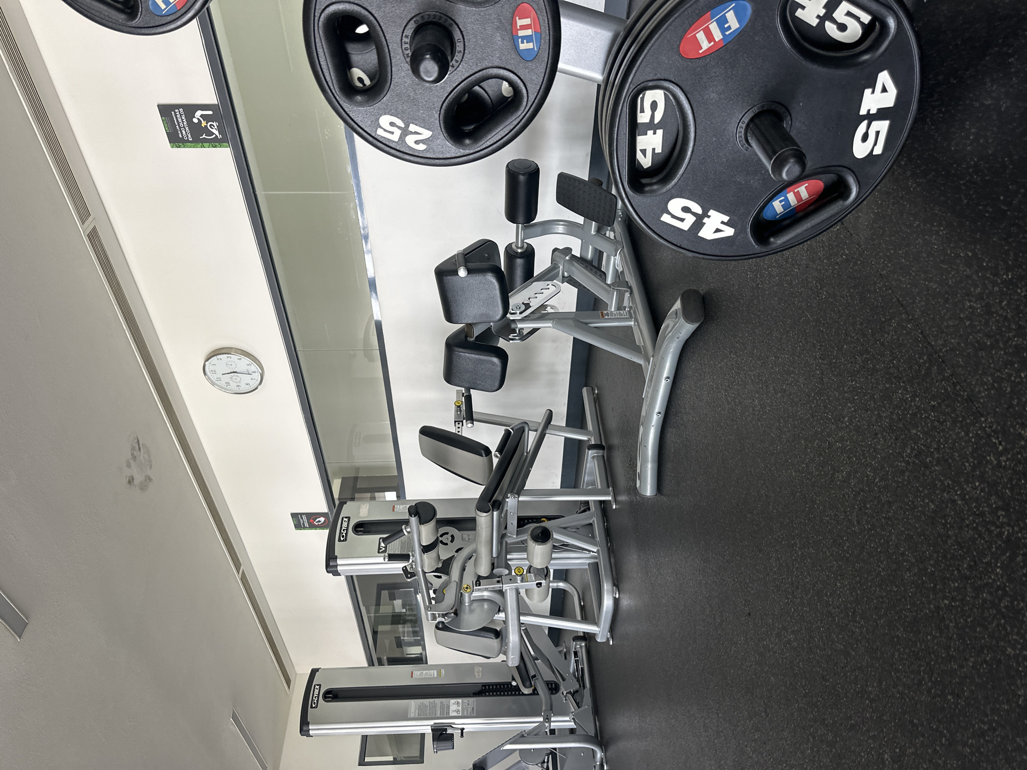 The image captures the interior of a modern fitness center, seen from an elevated perspective. The scene is devoid of people, presenting an orderly array of gym equipment.
Prominently featured are several black weight plates, clearly marked with white numbers "25" and "45," indicating their weight in pounds. Each weight plate also displays a distinctive red and blue "FIT" logo. Partially visible text on one plate includes "MEMBER" and "U.S. PAT. 052320."
Various strength training machines, identified by the "CYBEX" brand name, are arranged on a dark rubberized floor. These machines feature padded seats and grips, finished in silver and black. Light-colored walls surround the space, and reflective surfaces, possibly large mirrors or windows, run along one side, reflecting parts of the gym's interior.
Notable details include a wall clock showing approximately 9:15, and ventilation grates on the ceiling. A small green sign, partly reflected, features a wheelchair accessibility icon and the Spanish text "COMO QUIERAS ENCONTRARLOS" (meaning "However you want to find them"). The overall impression is that of a well-equipped and maintained gym, located in Huixquilucan de Degollado, Mexico, ready for use by members.
