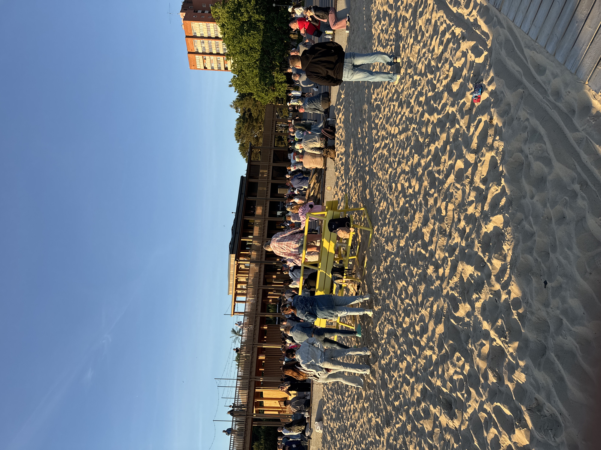 In Tallinn, Estonia, a sandy beach area adjacent to a wooden boardwalk hosts a gathering of people. The scene unfolds under a clear blue sky, with the warm, low-angle sunlight casting long shadows across the sand, suggesting it is late afternoon or early evening.
In the foreground, the sand is textured with ripples and footprints, indicating recent activity. A bright yellow metal structure, possibly a playground or seating area, is positioned on the sand. On this structure, several individuals are visible, including one person wearing a pink and white plaid shirt and shorts, and another in a purple hooded jacket.
Further back, a group of people are standing and sitting, many dressed casually in jeans and t-shirts, engaging in conversation or observing their surroundings. Behind them, a large, multi-level wooden structure with railings and open-air walkways dominates the mid-ground. This building appears to be a modern architectural feature, possibly a restaurant, bar, or event space, with lights strung across its upper levels.
In the background, a tall, orange brick apartment building stands out against the sky, suggesting the proximity of residential areas. Lush green trees flank the wooden structure and extend towards the building. A few individuals are visible on the upper levels of the wooden building, looking out at the scene. The overall impression is of a vibrant public space where people are enjoying the pleasant weather and social atmosphere.