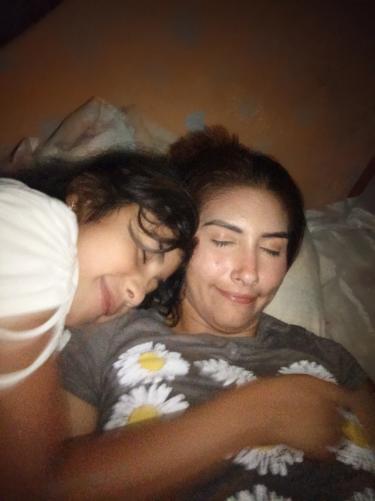A close-up, indoor shot shows a young girl and an adult woman lying down, apparently asleep or resting. The girl, on the left, has dark hair and is wearing a white shirt with thin straps. Her face is turned towards the camera with her eyes closed and a slight smile. The woman, on the right, has dark hair pulled back, a darker-colored shirt with a white floral print with yellow centers, and closed eyes, also with a gentle smile. They appear to be lying on a pillow or soft surface, with a white material (possibly bedding) visible behind them. The background is dimly lit and indistinct. There is no information in the image indicating that the location is Juan Griego, Venezuela.