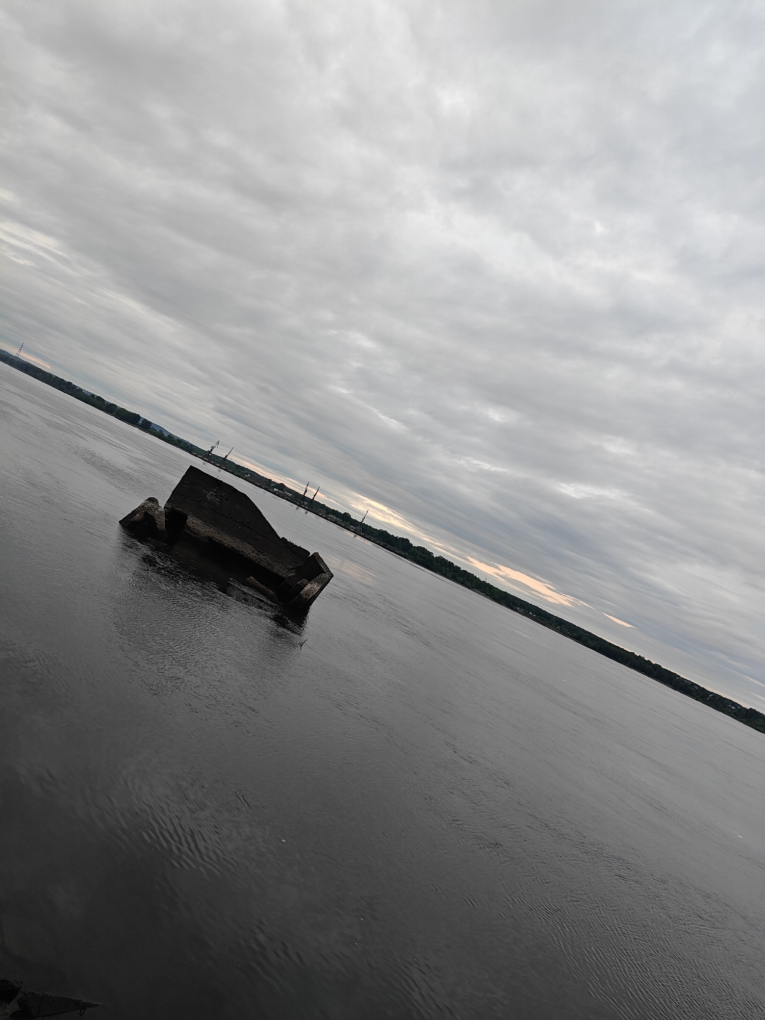 The image depicts an overcast waterscape in the city of Chaikovsky, Russia, under a heavily clouded sky. In the mid-ground, a large, dark, angular structure, possibly concrete or heavily weathered stone, is partially submerged in the calm, dark water. The structure shows signs of deterioration, with irregular surfaces and visible edges suggesting a remnant of a larger, possibly industrial, installation. The water exhibits subtle ripples across its surface. In the distant background, a tree-lined shoreline is visible, extending along both sides of the expansive body of water. Along the right distant shore, multiple industrial cranes and associated structures characteristic of a port or heavy industry are discernible. The sky is uniformly covered with dense, grey clouds, with only faint hints of lighter tones near the horizon on the right side. No individuals or animals are present. The photograph is taken at a steep, diagonal angle.