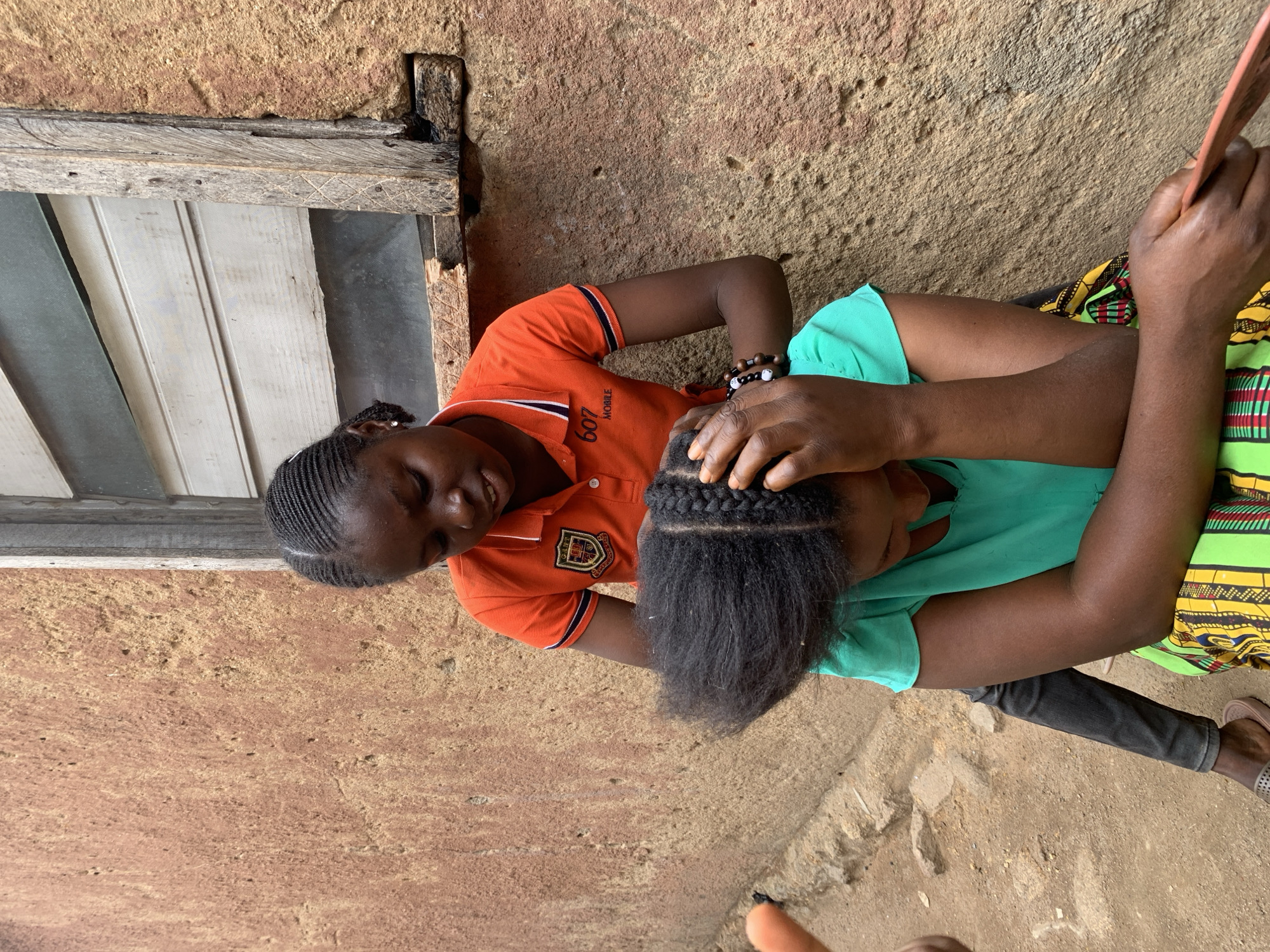 Two Black African women are sitting outdoors against a textured, earthen-colored wall with a weathered wooden window frame visible to the left. The woman on the left wears an orange polo shirt and has her hair neatly braided. She is facing the woman on the right, whose hair is partially braided with the rest unbraided. The woman in orange appears to be working on the hair of the other woman. The woman on the right is wearing a light green shirt and dark pants. A colorful patterned fabric is visible under her. This scene is located in Jos, Nigeria.