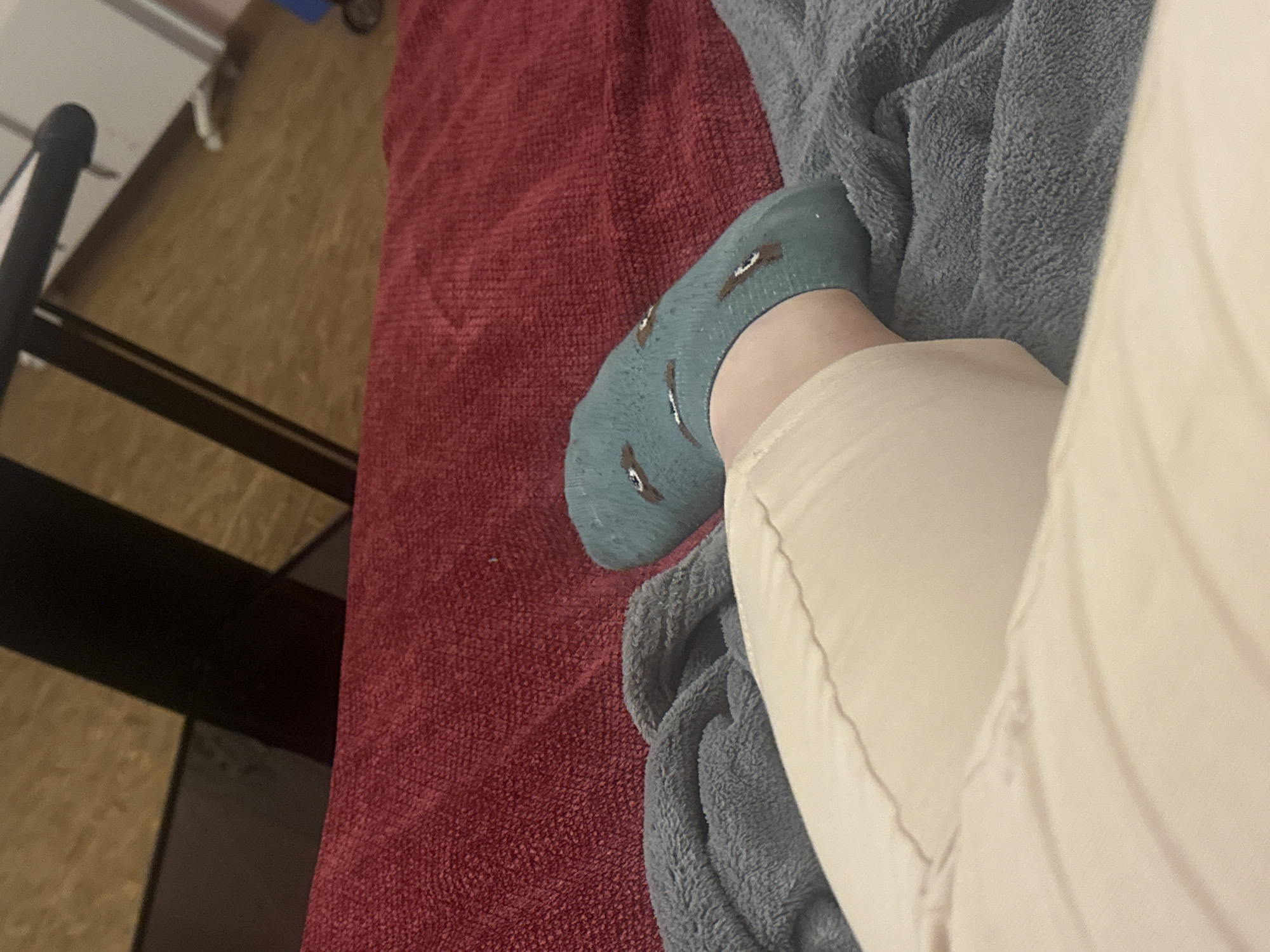 A close-up, low-angle shot shows a person's leg and foot. The foot wears a turquoise sock with a pattern of small, brown objects. The leg is covered by tan-colored pants or pajamas. The leg rests on a light gray fleece blanket. Below the blanket is a dark red, textured blanket. To the left of the blankets is a dark brown and black shelving unit and light brown flooring. The setting does not appear to be in Razhevo Konare, Bulgaria. There are no indications of the location from the image.