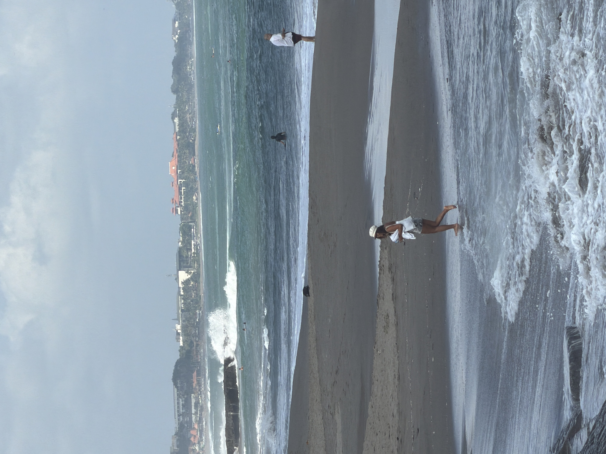 This is a photograph taken on a beach in North Kuta, Indonesia, with a cloudy sky overhead. In the foreground, a woman wearing a white hat and a patterned skirt is walking near the water's edge, carrying a white bag. The sand is dark and wet, with gentle waves lapping the shore. In the middle ground, a man in a white shirt and dark shorts stands on the wet sand, facing the ocean. Further out in the water, a person is seen with a surfboard. The background reveals a coastline with buildings, some with red roofs, nestled amongst palm trees, indicative of a resort area. The ocean is choppy with visible waves, suggesting a moderately active surf.