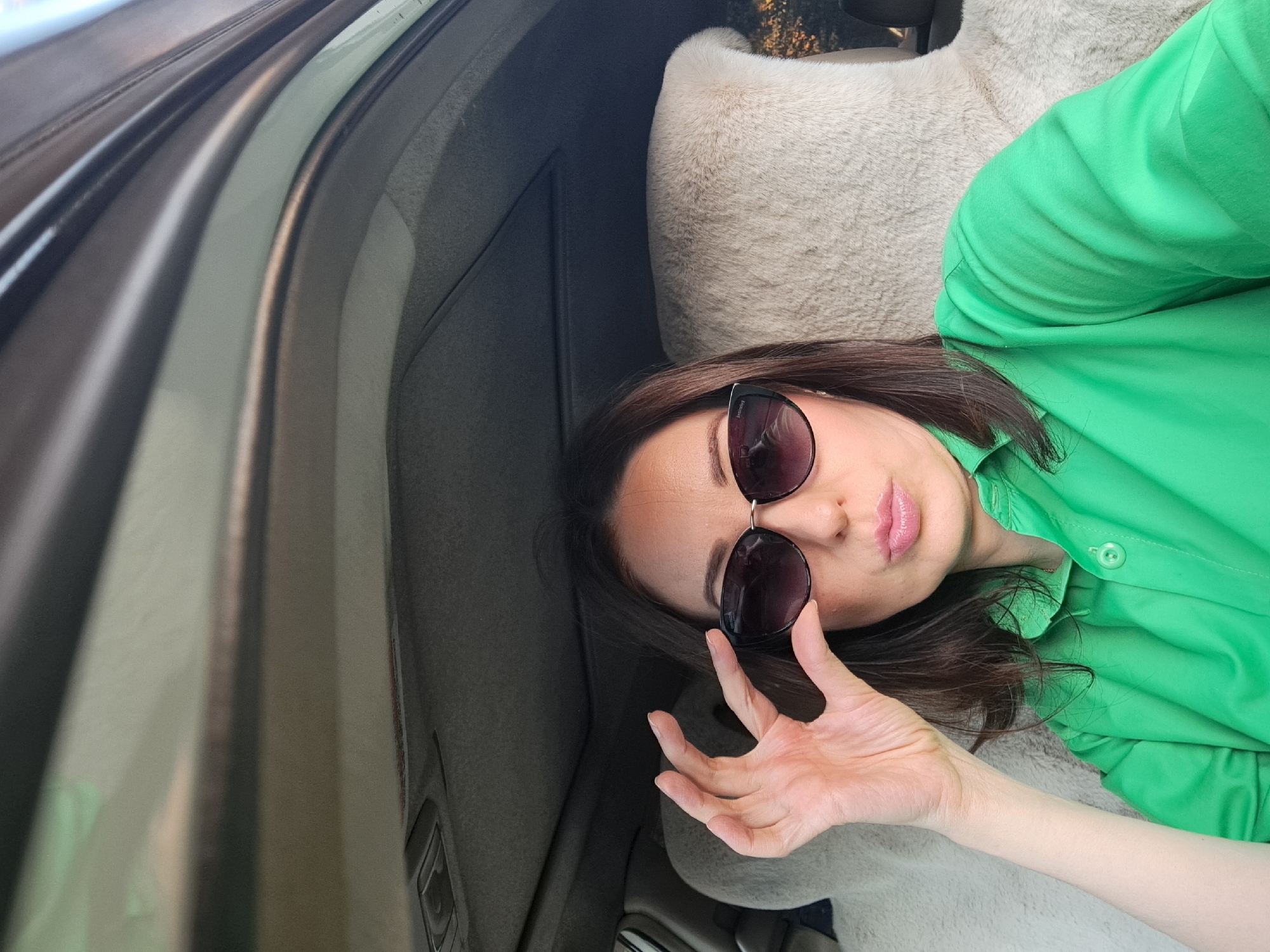 A woman is sitting in the passenger seat of a car, looking at the camera. She is wearing a bright green collared shirt and dark sunglasses. Her left hand is raised to adjust her sunglasses. She has dark brown hair and is wearing light pink lipstick. The interior of the car is visible, including the headliner, sunroof, and the fuzzy sheepskin cover on the passenger seat. The background outside the car suggests an outdoor, possibly wooded area. There is no identifiable information to place this image in Irkutsk, Russia.