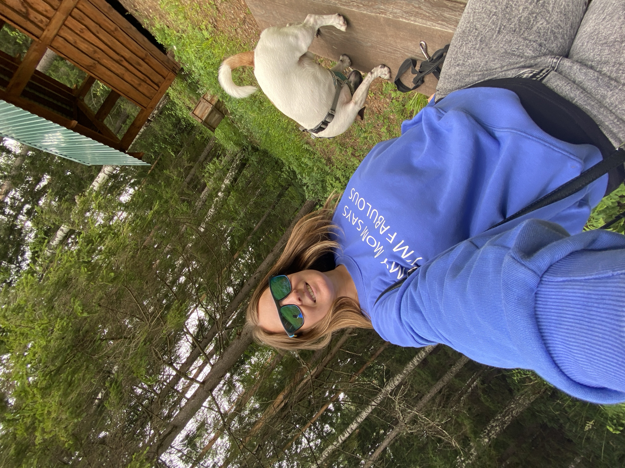 In Noviy, Russia, a person is taking a selfie outdoors, likely during the daytime. The individual is wearing a blue sweatshirt with white text that reads "MY MOM SAYS I'M FABULOUS" and grey jeans. They are also wearing sunglasses. The scene is set in a wooded area, with tall trees and lush green grass visible. To the left of the frame, there's a rustic wooden structure with a corrugated metal roof. A white dog with a tan tail is lying on a wooden bench in the background, wearing a harness. The dog appears relaxed. The overall mood of the image is casual and happy, with the person smiling at the camera. The weather appears to be pleasant, given the outdoor setting and the person's attire.