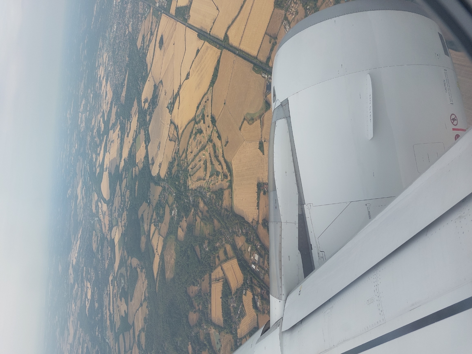 An aerial view captured from an airplane window shows a landscape of rural farmland and scattered settlements under a hazy sky. The foreground is dominated by the white, metallic casing of an airplane engine and a portion of the wing. Beyond the aircraft components, a patchwork of golden-brown fields, darker green wooded areas, and small clusters of buildings stretches towards the horizon. A road or railway line bisects the landscape in the middle distance. The image is a still, observational shot, typical of an in-flight photograph, and offers no discernible human activity or interaction. The specific geographical location within Ruscombe, United Kingdom, cannot be pinpointed solely from the visual information provided in the image.