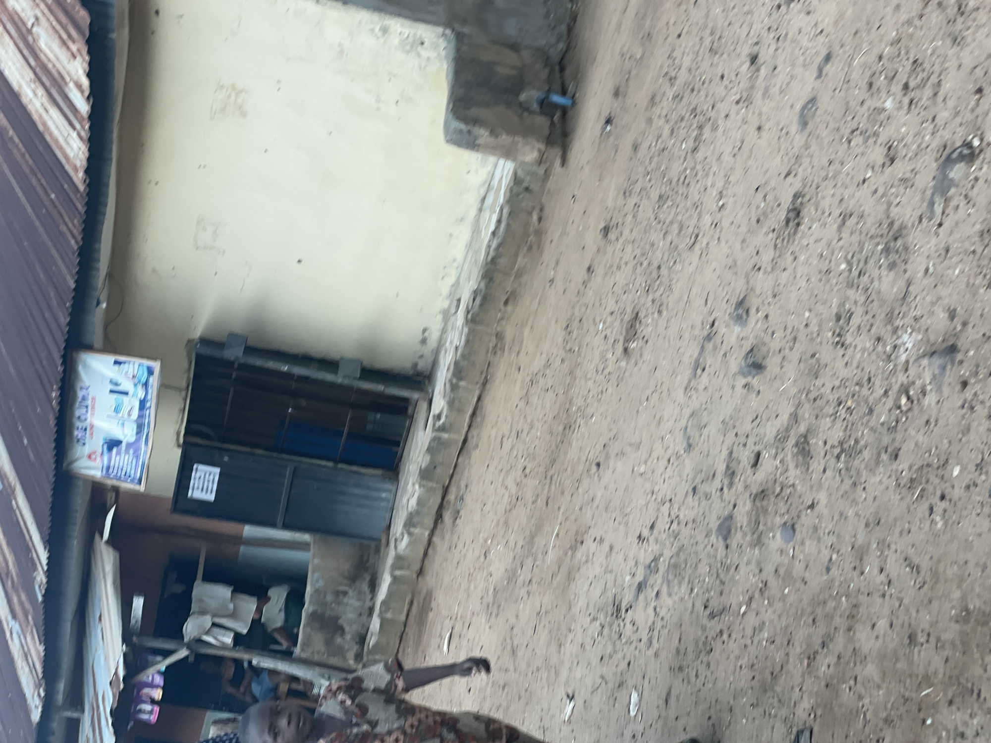 The image depicts an outdoor scene in Lokoja, Nigeria, likely a street in a commercial or residential area. In the foreground, a young boy wearing a patterned shirt walks towards the viewer. The ground is unpaved, composed of sand and small stones.
Behind the boy, there is a building with a corrugated iron roof. Attached to the building is a covered stall area with items displayed on shelves. A sign is visible above the stall, showing product images and text in an unknown language. Adjacent to the stall is a metal-framed doorway leading into a darker interior space, with bars or a gate visible.
Further to the right, the building has a pale yellow-colored wall. There is a concrete ledge or raised sidewalk running along the front of the buildings. In the background, on the right side of the image, there's a concrete structure and a blue pipe. The overall impression is of a functional, possibly developing, urban environment.