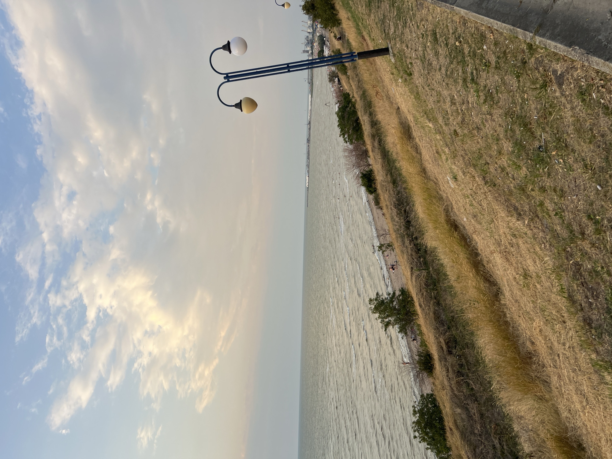 The image depicts a coastal view at Yeysk, Russia, featuring a body of water, a beach, and a grassy hillside. The sea is calm with gentle waves, meeting a sandy beach where a few people are visible. The hillside is covered in dry, golden grass and sparse vegetation, sloping downwards towards the beach. A distinctive blue lamppost with two lights stands on the right side of the frame, on the upper part of the hillside. In the distance, along the coastline, industrial structures and buildings can be observed, suggesting a port or urban area. The sky is partly cloudy, with a soft, diffused light indicating either early morning or late afternoon.