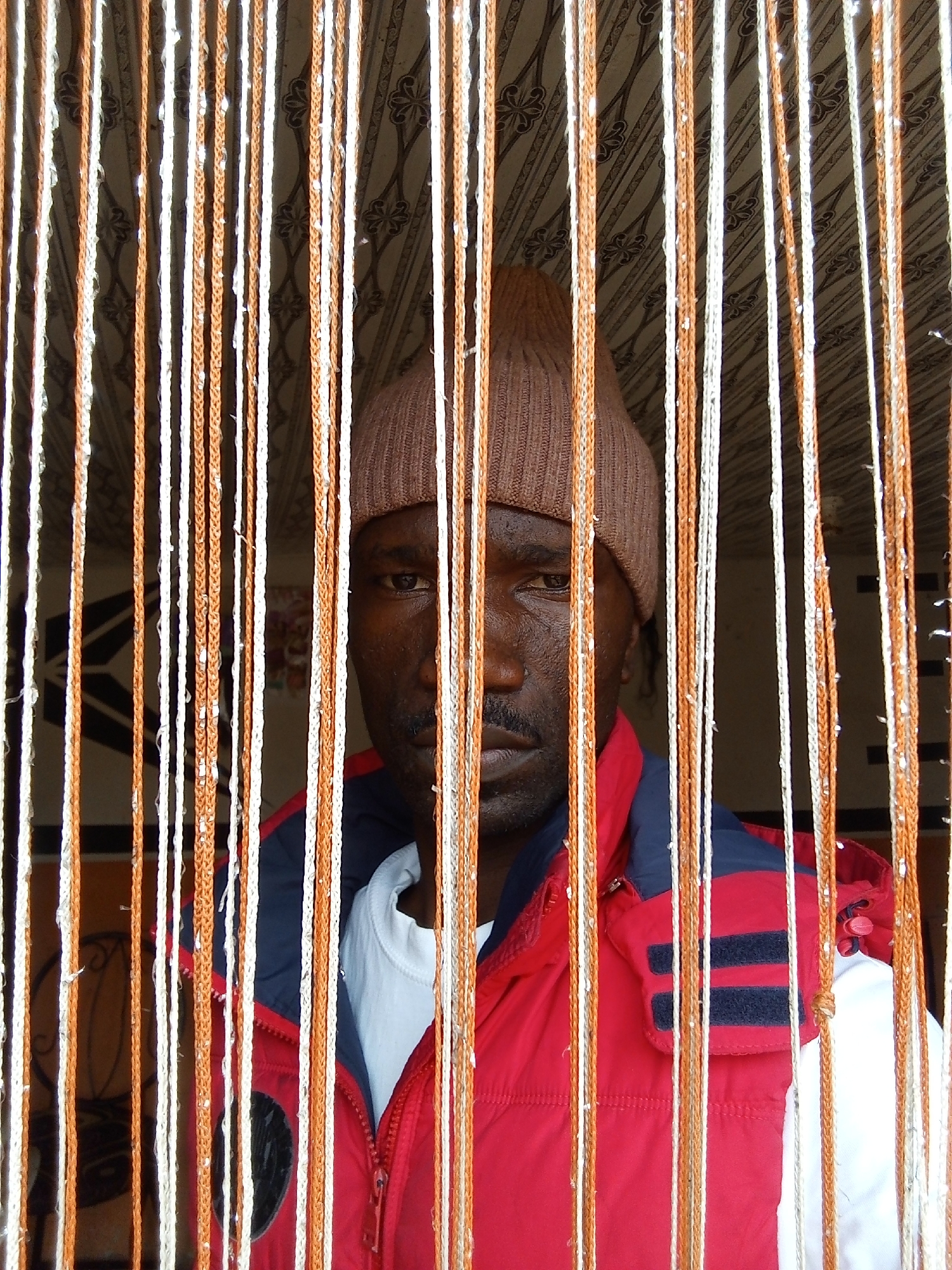 A medium shot reveals a man standing behind a curtain of vertically oriented strands of white and orange thread. He wears a brown knit cap and a red jacket with navy blue accents over a white shirt. The background, visible through the curtain, shows a patterned ceiling or wall covering. The scene is likely indoors, but the specific location within the city of Jos, Nigeria, is not discernible from the image. There are no discernible actions or interactions occurring in the image.
