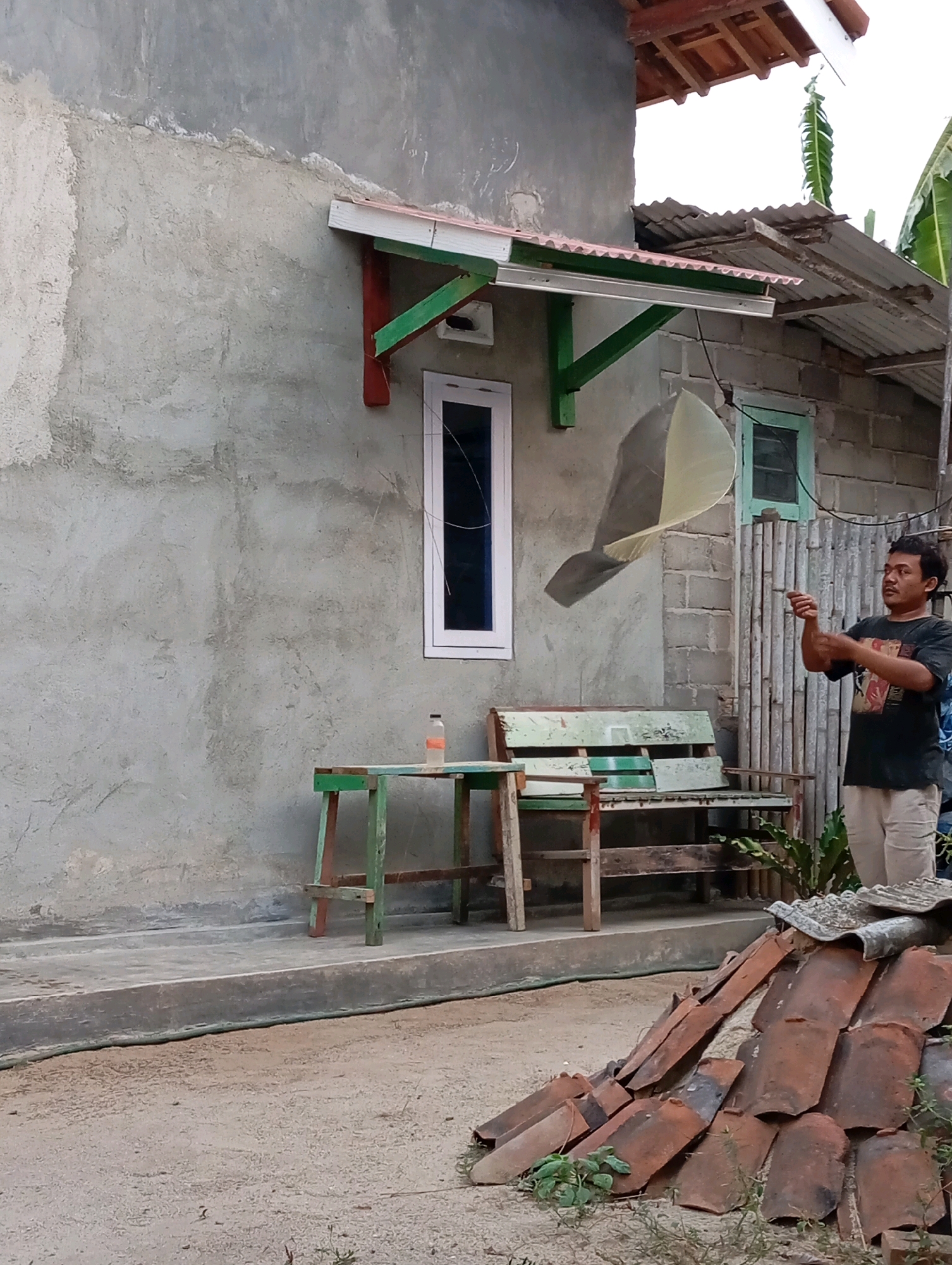 A young man stands to the right of a weathered, gray concrete wall, looking upwards and to the left. He is wearing a dark t-shirt and light-colored pants. In his hands, he holds a string, and he appears to be flying a kite, which is visible in the upper left quadrant of the image, caught in the wind. In front of the wall, a light green wooden bench with a slatted seat and backrest sits on a concrete walkway. To the left of the bench is a green and red wooden table with a bottle on it. In the foreground, a pile of reddish-brown roof tiles lies on a sandy ground. The background shows a corrugated metal roof and some green vegetation. The scene is set in Leman Benawi, Indonesia, a rural or semi-rural area.