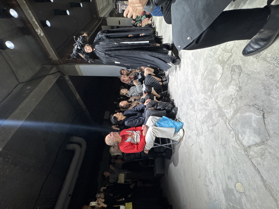 The image depicts an indoor fashion runway show taking place in a raw, industrial-style venue in Paris, France. A model is observed walking from left to right on a concrete runway, illuminated by overhead spotlights. The model wears a voluminous, multi-layered black garment featuring a long, flowing silhouette and an elaborate, sculptural black headpiece with organic forms, paired with substantial black platform boots.
To the left of the runway, rows of spectators are seated on simple folding chairs. Many audience members are holding smartphones elevated, actively recording the event. A prominent male spectator in the foreground, seated in the front row, is bald and wears a red long-sleeved shirt with a graphic design, light-colored trousers, and white sneakers. A blue plastic bag rests on the floor beside his right leg. Other attendees are dressed in a variety of contemporary attire, predominantly in dark or muted colors.
The venue's environment features exposed concrete ceilings with visible pipes and structural beams. A distinct shaft of light from an unseen source high above illuminates a portion of the ceiling on the upper left. The floor is rough concrete, exhibiting cracks and areas of discoloration. The overall scene is characterized by focused attention on the model's presentation, consistent with a high-fashion event.