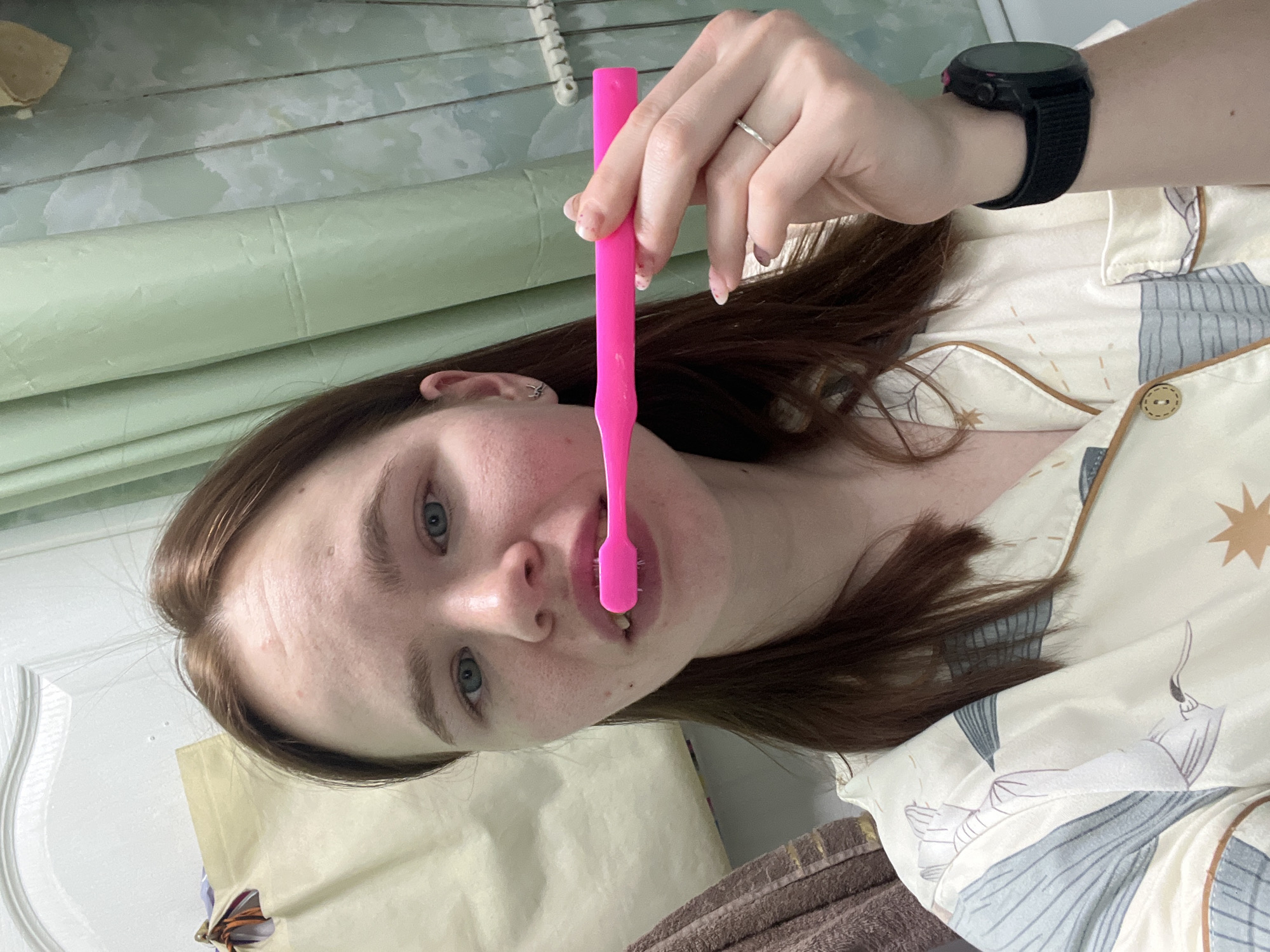 A woman is holding a pink toothbrush in front of her mouth. She is wearing a black watch on her left wrist and a ring on her left ring finger. She is wearing pajamas with a pattern of stars and figures. The background includes a green window covering and a white door. The location is not identifiable as Chaikovsky, Russia.